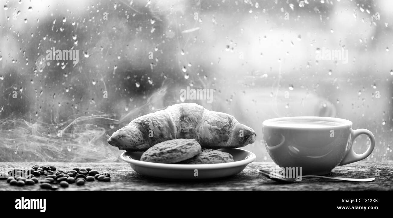 Frisch gebrühter Kaffee in weiße Tasse oder Becher auf der Fensterbank. Trinken Kaffee mit Croissant Dessert. Genießen Kaffee an regnerischen Tag. Kaffee Zeit an regnerischen Tag. Nasses Glas Fenster und heiße Tasse Koffein trinken. Stockfoto