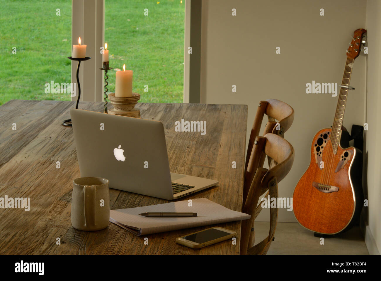 Arbeiten von zu Hause aus. Ein rustikaler Holztisch mit einem Laptop, Kerzen, einen schreibblock füllen Sie die Szene. Eine Ovation Gitarre lehnt sich gegen die Wand. Stockfoto