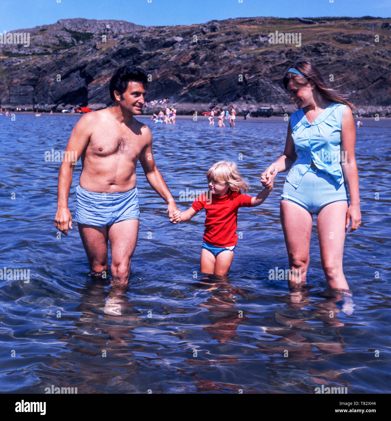 Ein Onkel und Tante mit einem jungen Mädchen. das Waten im Meer am schwarzen Felsen Sand. North Wales. Großbritannien. Stockfoto