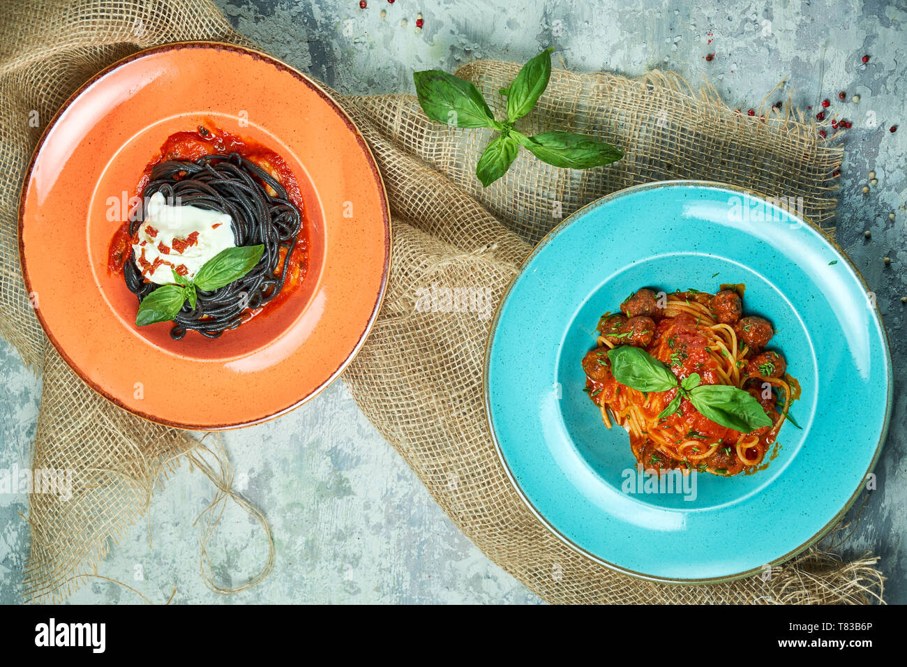 Blau Teller mit Nudeln und Fleisch Kugeln, orange Platte mit Tinte vom Tintenfisch Tinte. Graues Licht strukturierten Hintergrund. Schöne Servieren von Speisen. Restaurant Menü Stockfoto