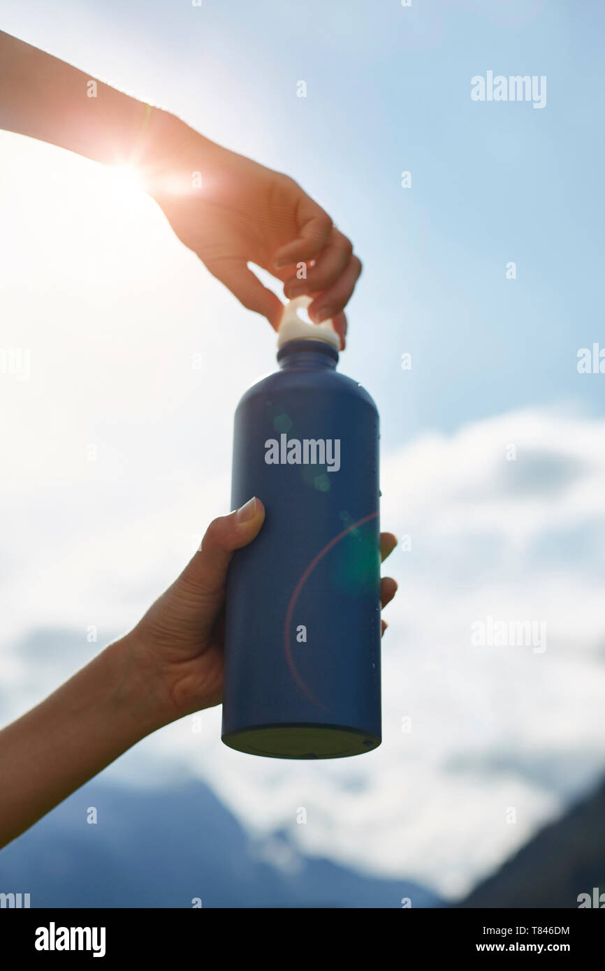 Weibliche Wanderer Eröffnung getränke Flasche gegen sonnendurchfluteten Himmel, 7/8-Ansicht der Hände Stockfoto