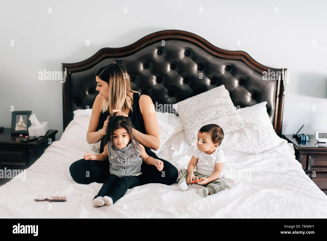 Baby Junge sitzt auf dem Bett, während die Mutter Stile Schwester Haar Stockfoto