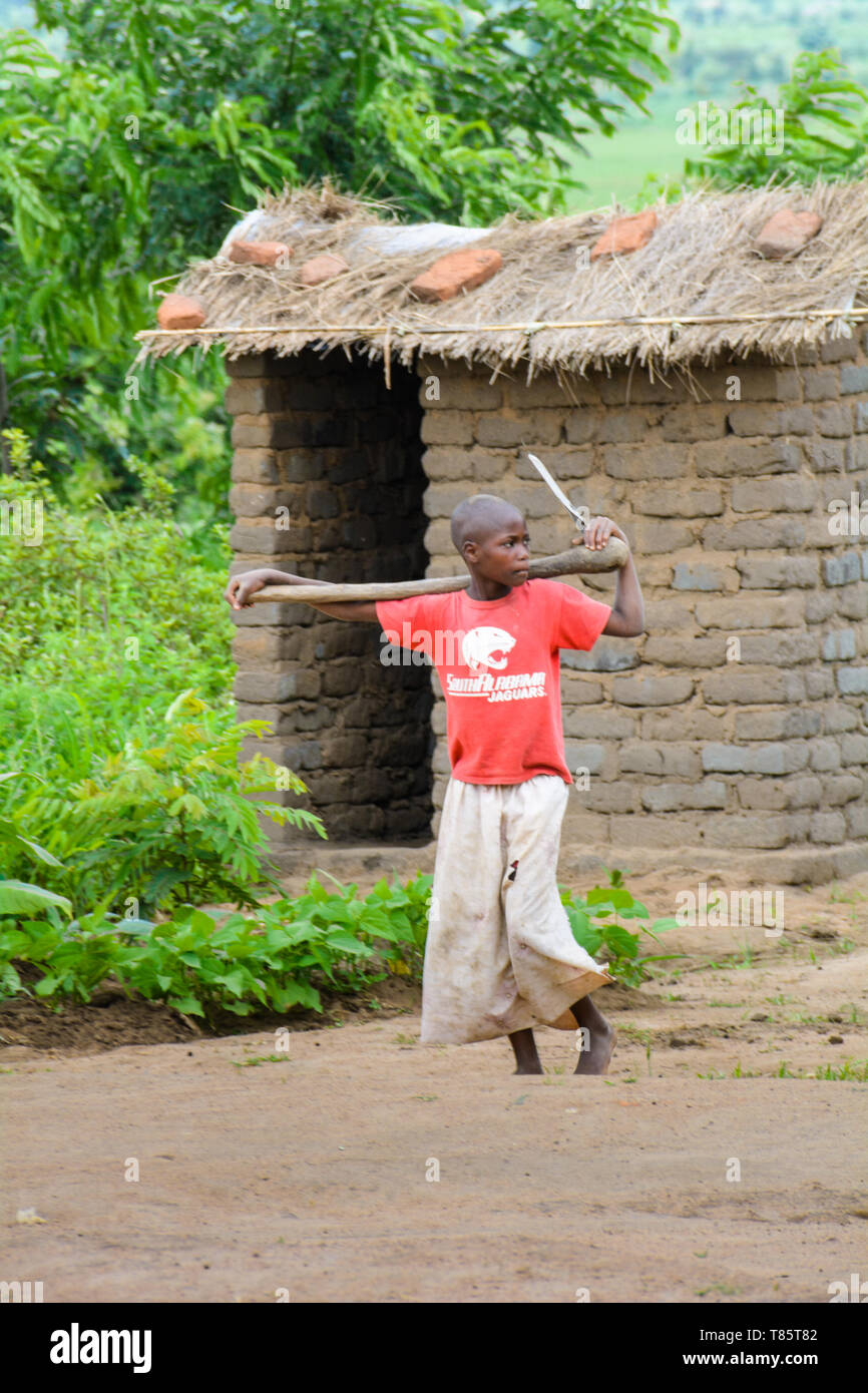 Barfuß malawische Mädchen gehen mit einem traditionellen Hacke auf ihren Schultern hinter einer traditionellen Latrine mit dem Gras Strohdach Stockfoto