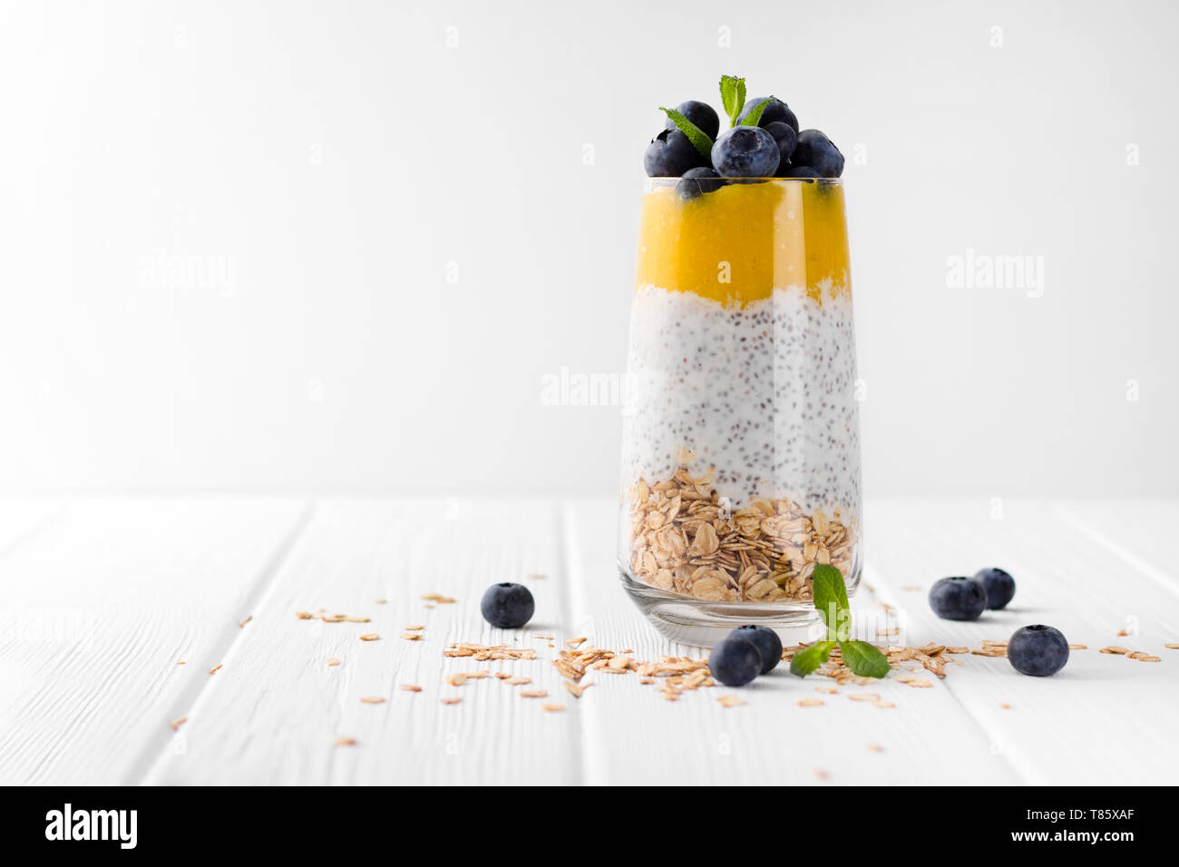 Glas Joghurt mit Chia Samen, Blaubeeren und Mango Mousse und Haferflocken auf einem weißen Holztisch. Stockfoto