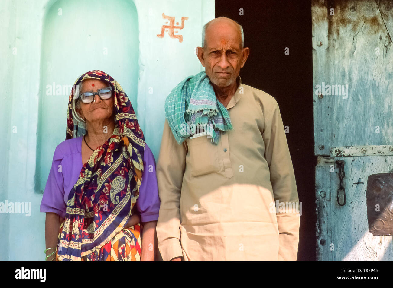 Porträt einer indischen Ehepaar im Alter, traditionelle Kleidung, an der Tür stehend vor ihrem Haus, religiöses Symbol an die Wand gemalt, Indi Stockfoto