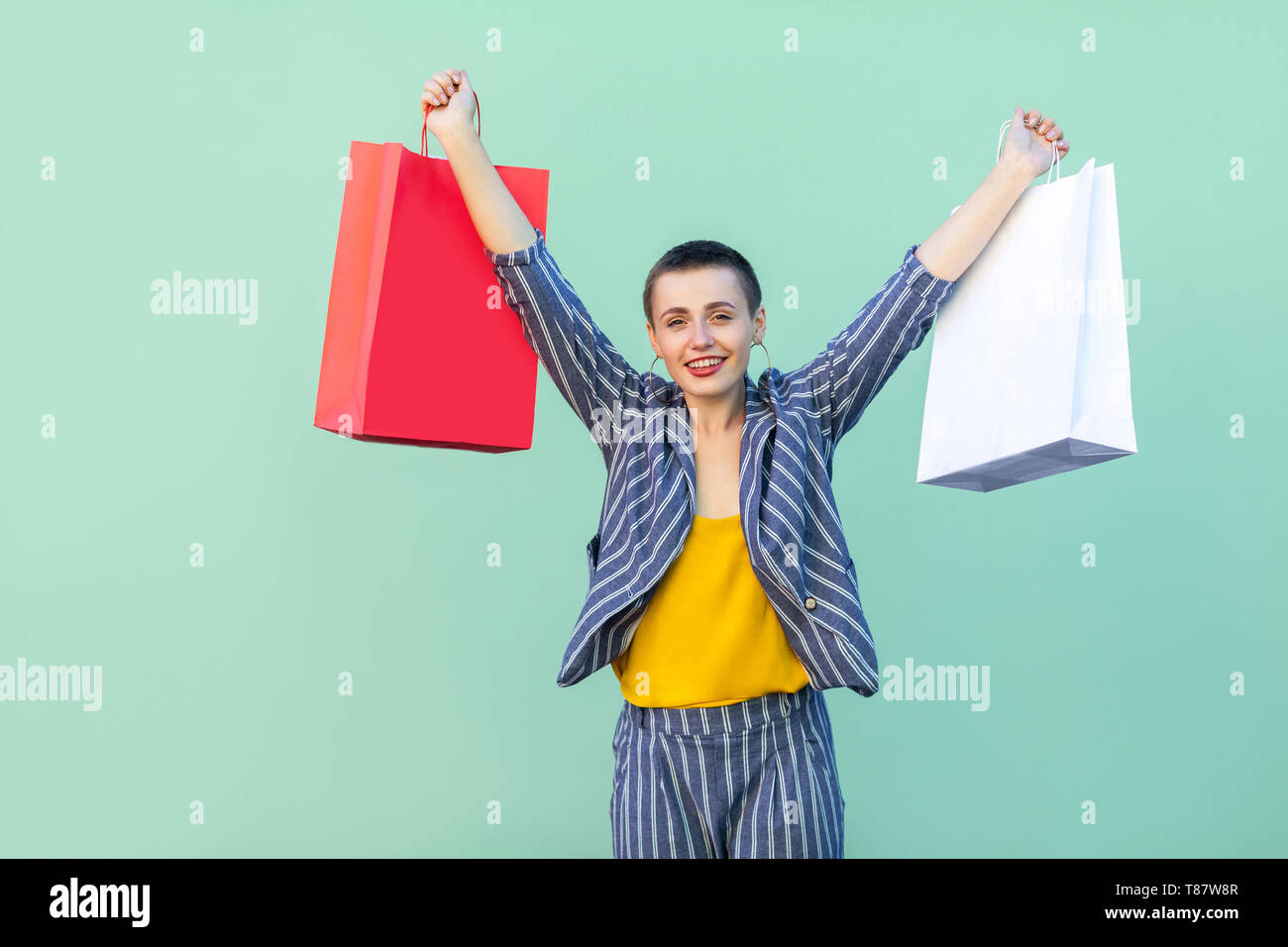 Schöne mit kurzen Haaren junge Frau im gestreiften Anzug stand Zufrieden, holding Einkaufstaschen mit erhobenen Armen, toothy Lächeln und Kamera. Stockfoto