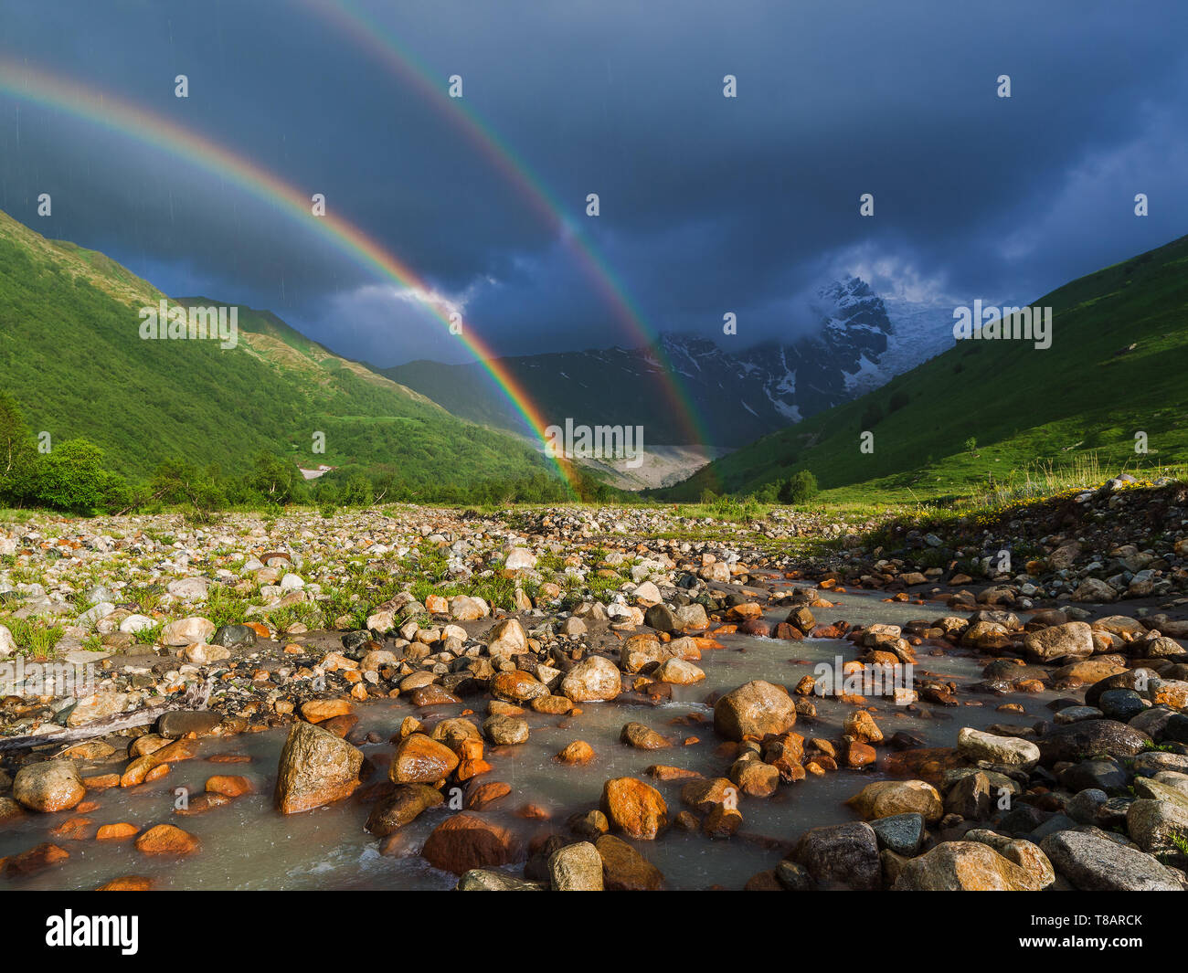 Regenbogen im Gebirge. Sommer Landschaft mit Fluss, Hügeln und einem stürmischen Himmel. Regen im Tal. Stockfoto