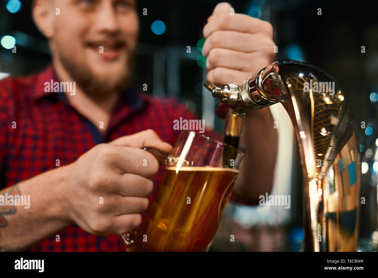 Selektiver Fokus der männlichen Händen Pint und Gießen Bier in der Kneipe. Junge Bartgeier Barkeeper in kariertem Hemd mit Tattoo arbeiten und warten die Menschen in der Bar. Konzept der Ale und Job. Stockfoto