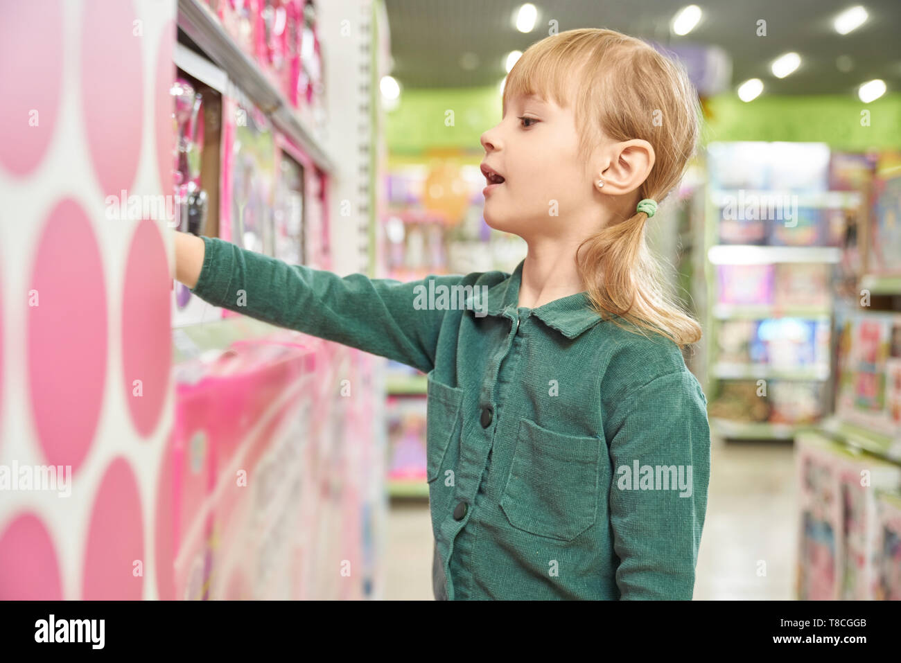 Kleines, hübsches Mädchen Auswahl Spielzeug in großen Einkaufszentrums. Fröhliche erstaunt Kind mit geöffnetem Mund in der Nähe von Regalen mit Kindern Spielzeug. Junge Kunden die beste Spielzeug. Stockfoto