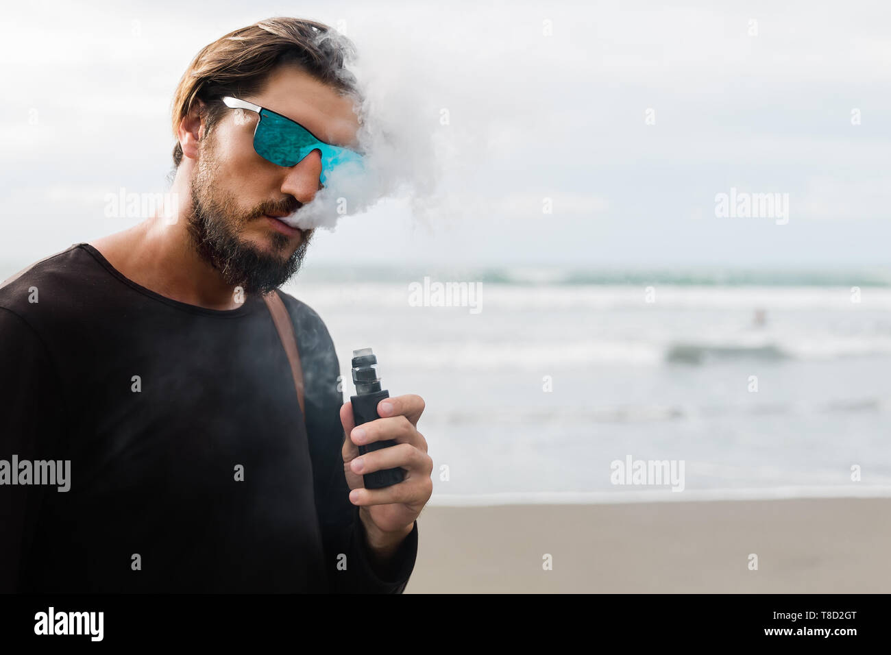 Mann mit Bart und Sonnenbrille rauchen Elektronische sigarette Outdoor am Sunset Beach. Junge männliche Hipster vaping am Meer. Stockfoto