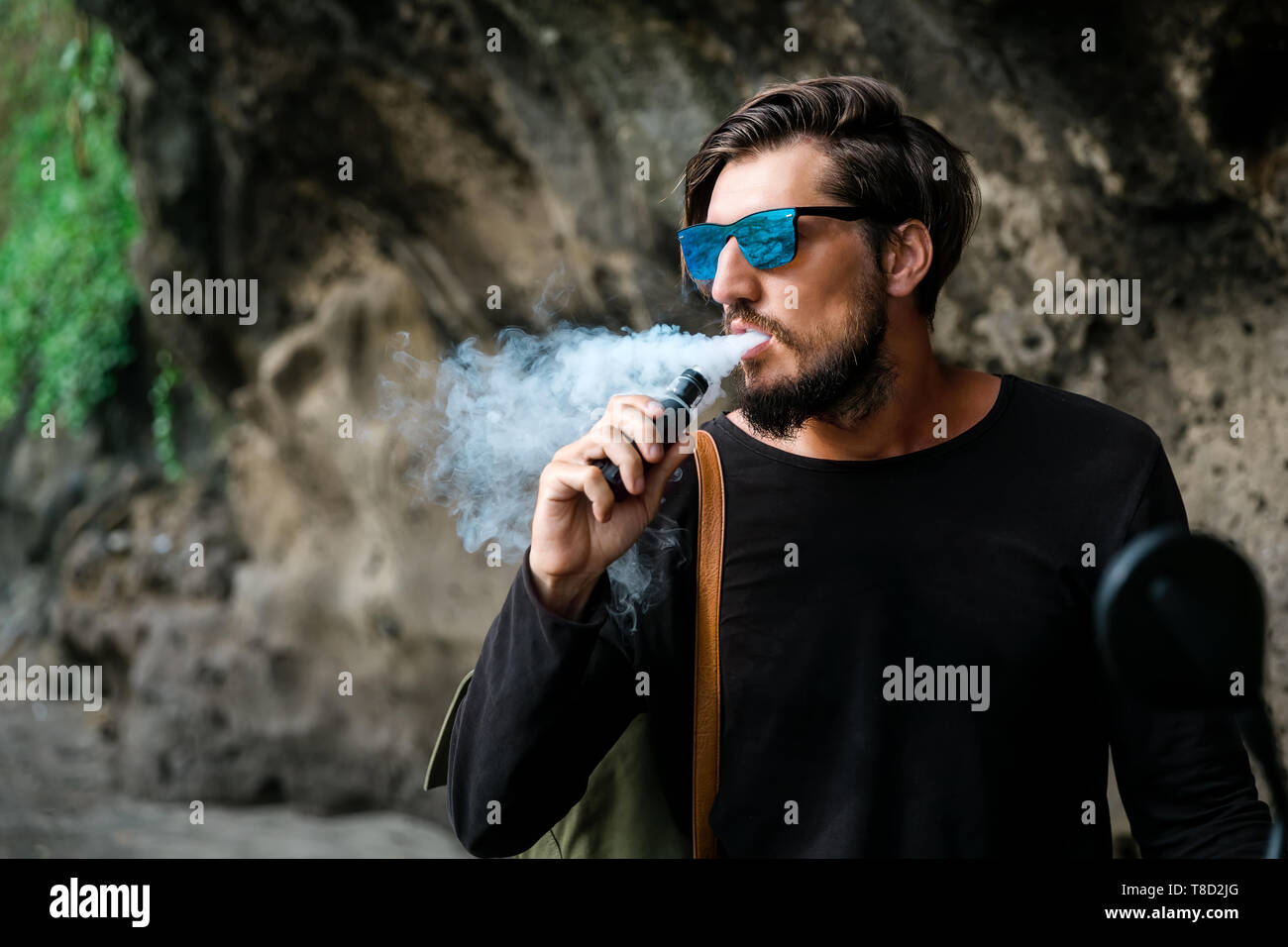Mann mit Bart und Sonnenbrille Sitz auf Motorrad rauchen Elektronische sigarette Outdoor am Sunset Beach. Junge männliche Hipster vaping am Meer. Stockfoto