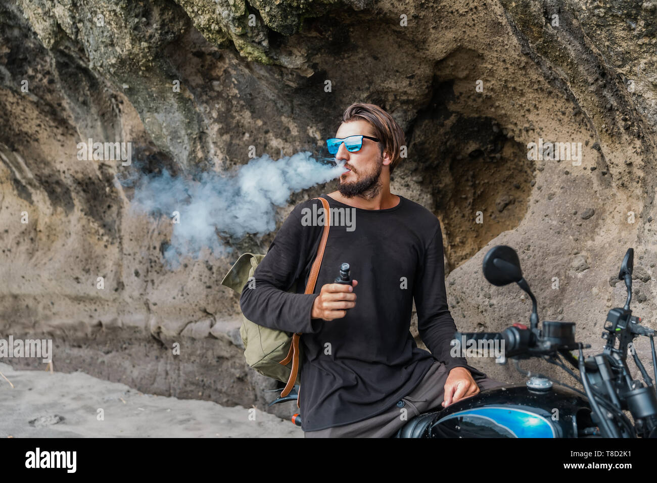 Mann mit Bart und Sonnenbrille Sitz auf Motorrad rauchen Elektronische sigarette Outdoor am Sunset Beach. Junge männliche Hipster vaping am Meer. Stockfoto