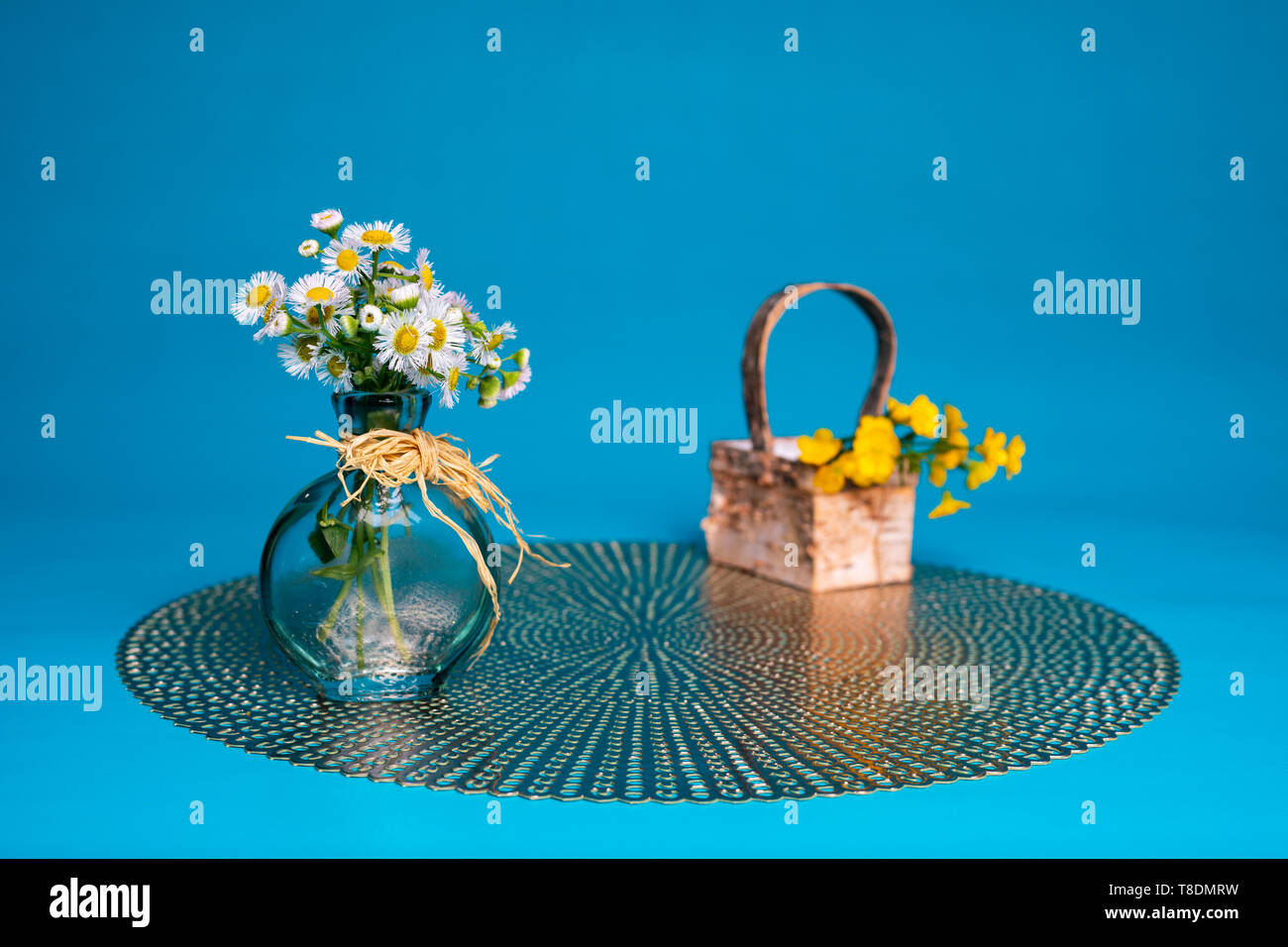 Prairie fleabane Wildblumen in ein kleines Glas Vase gegen eine hellblaue Hintergrund angeordnet. Ein paar haarige buttercup Blumen liegen in einem kleinen Korb. Stockfoto