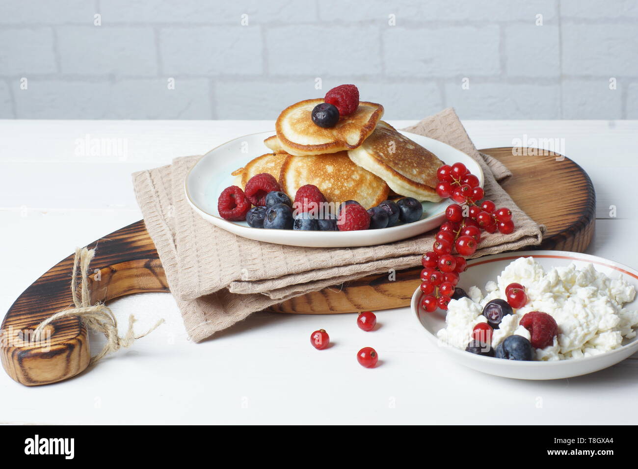 Auf dem Tisch liegt ein Brett mit Pfannkuchen mit frischen Beeren der Johannisbeere, Heidelbeere und Himbeere, und neben einer Schüssel Quark Stockfoto