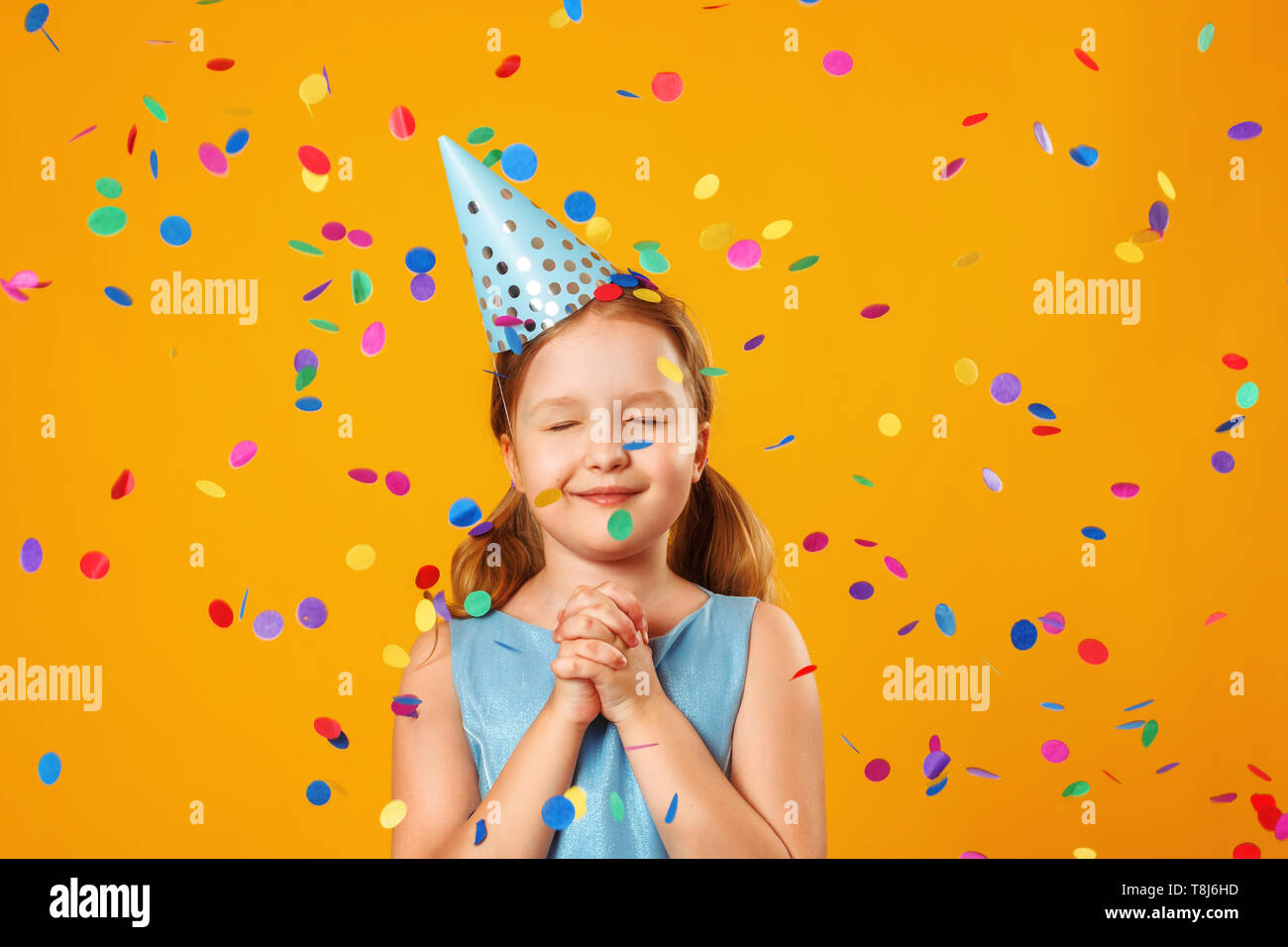 Süße kleine Mädchen feiert Geburtstag. Das Kind schloss die Augen und macht einen Wunsch im Regen aus Konfetti. Closeup Portrait auf gelben Hintergrund. Stockfoto