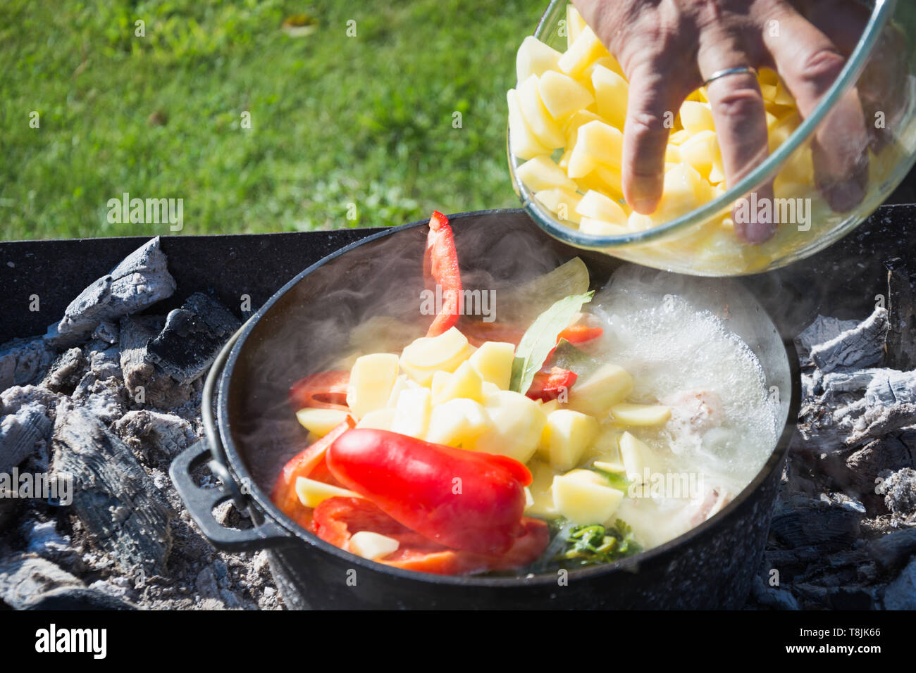 Shurpa Suppe in einer großen gusseisernen Kessel outdoor. Sommer essen für Wanderung. Das Kochen. Türkische Küche. Mens hand Zutaten für die Suppe. Stockfoto