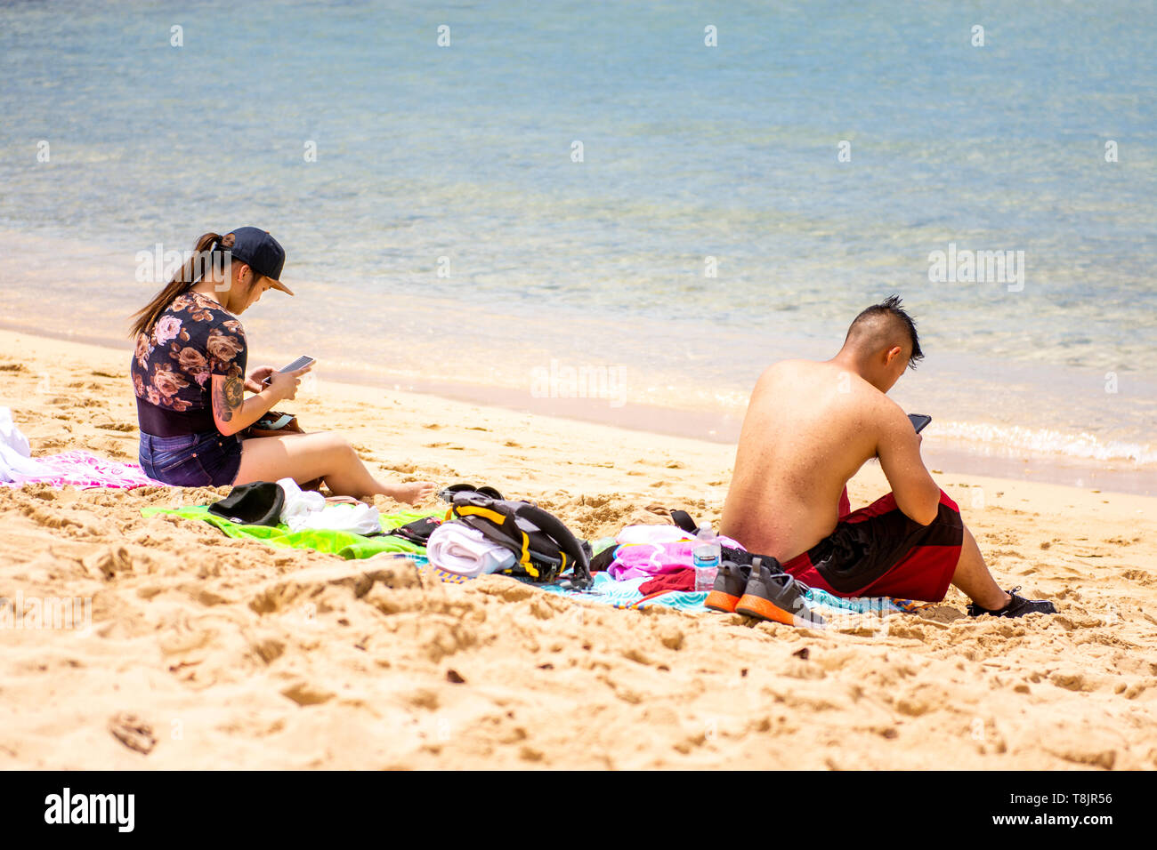 Zwei Teenager, Asiaten an einem Strand in Hawaii, beide haben ihre Telefone angeschlossen. Stockfoto