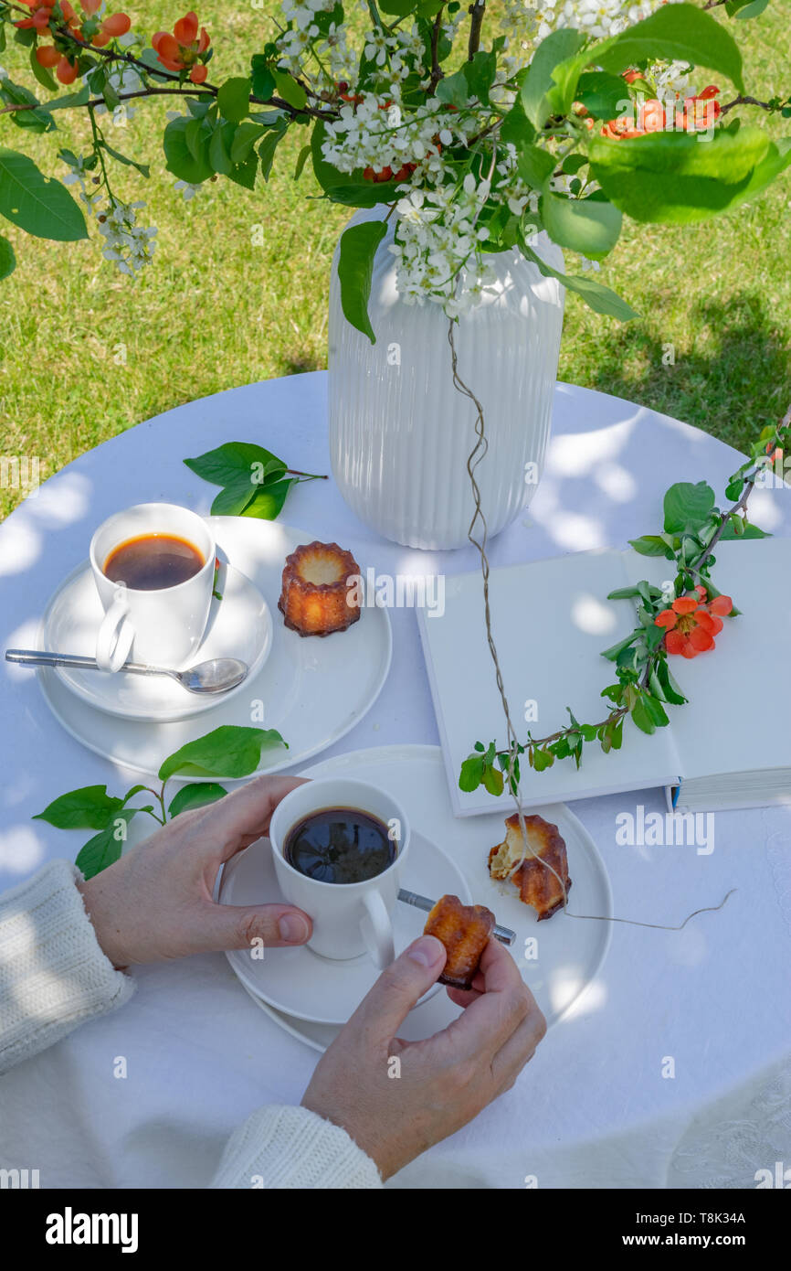 Genießen Kaffee Zeit im Garten. Serviert mit Kaffee, canele, Vase, Blumen und Buch im Garten. Anordnung mit den Händen einer Frau. Stockfoto