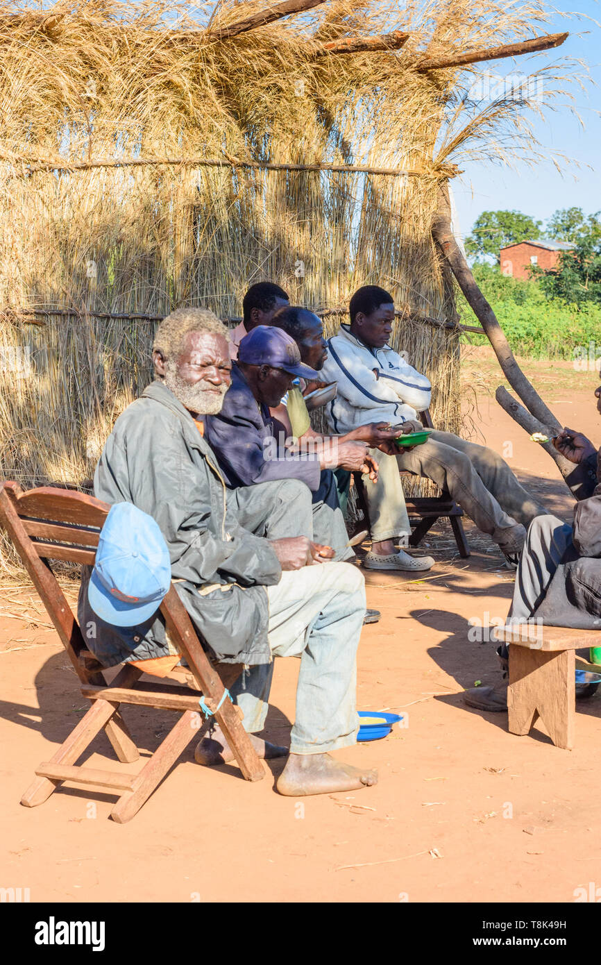 Ein alter Mann ruht auf einem tradtionally gebaut Holzstuhl essen ein Frühstück von Mais porridge unter anderen Männern in Nsanje, Malawi Stockfoto