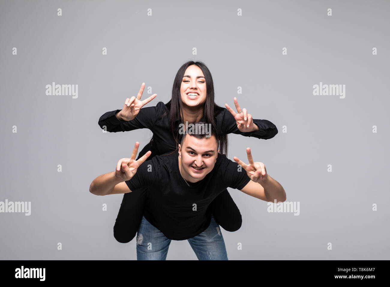 Junge lustig Paar, Freund piggy backing Freundin, Mädchen, Frieden Zeichen mit den Händen über grauer Hintergrund Stockfoto