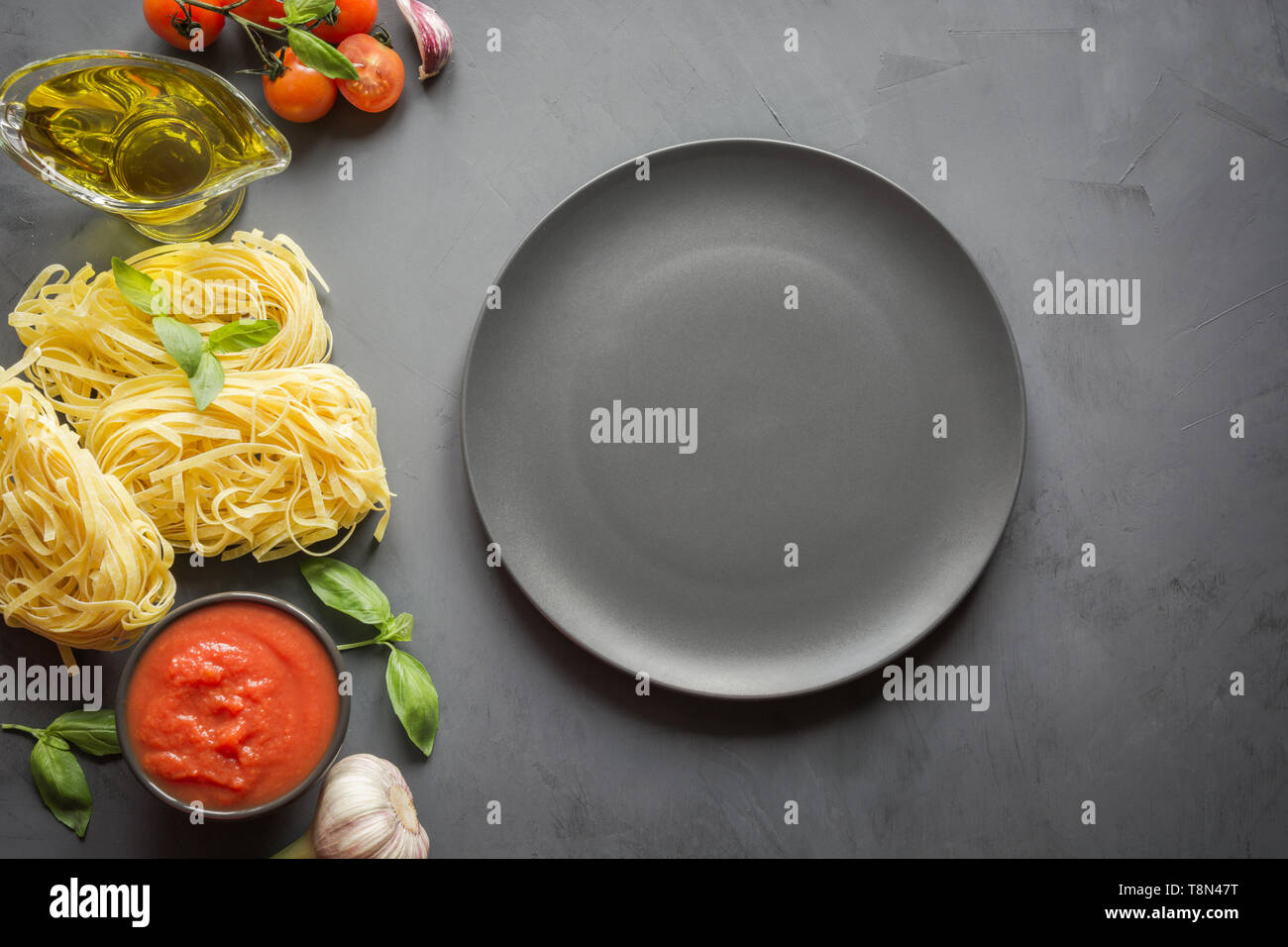 Leere schwarze Platte und Grenze der rohen Pasta, Tomaten im eigenen Saft, Basilikum, Parmesan zum Kochen italienische und mediterrane Gerichte auf grauem Beton Stockfoto