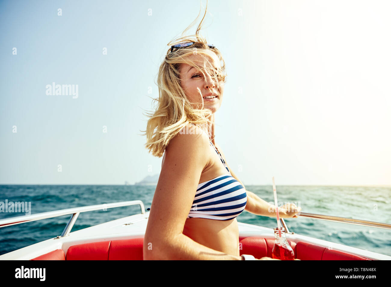 Lächelnde junge Frau im Bikini Top Entspannung auf einem Boot mit einem Getränk an einem sonnigen Tag während ihrer Sommerferien Stockfoto