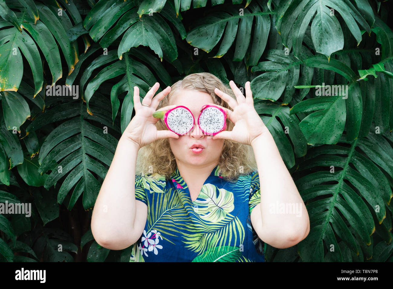 Blonde Caucasian woman holding Scheiben Drachenfrucht Pitaya der vor ihren Augen, küssen. Tropische Blätter Hintergrund, kopieren. Stockfoto