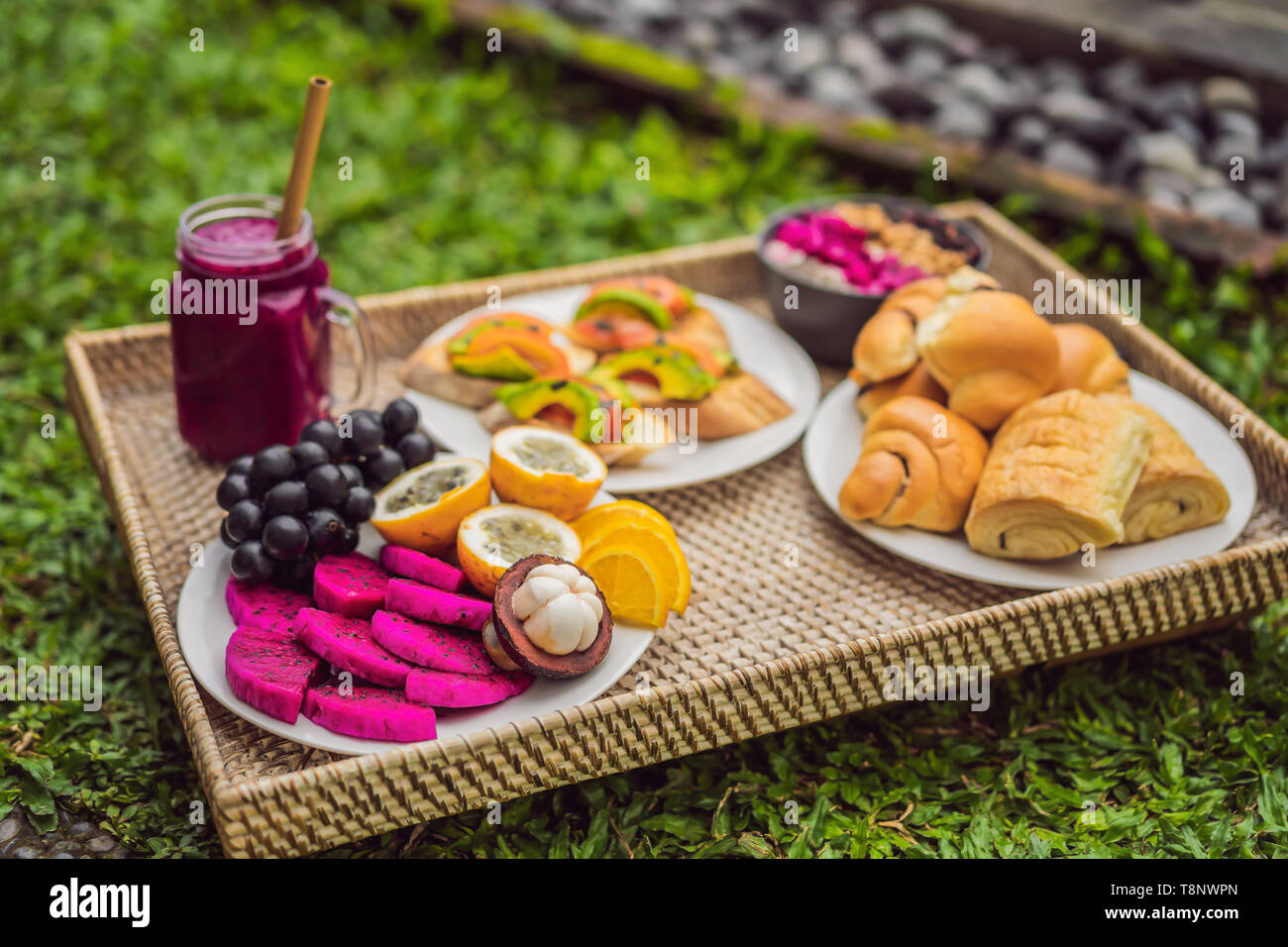 Frühstück auf einem Tablett mit Obst, Brötchen, Avocado Sandwiches, smoothie Schüssel stehen auf dem Gras Stockfoto