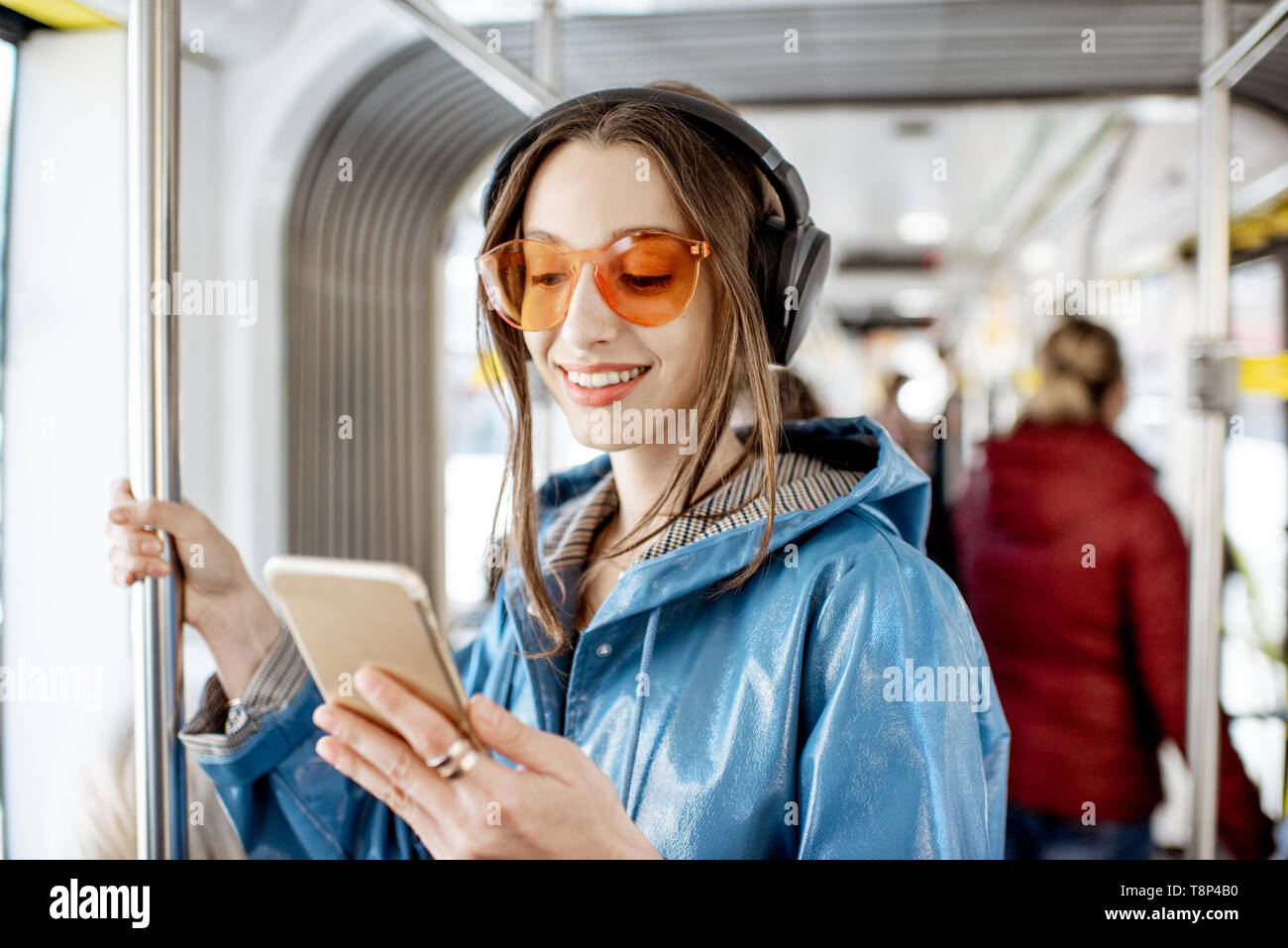 Junge elegante Frau mit öffentlichen Verkehrsmitteln, stehend mit Kopfhörern und Smartphone, während in der modernen Straßenbahn verschieben Stockfoto