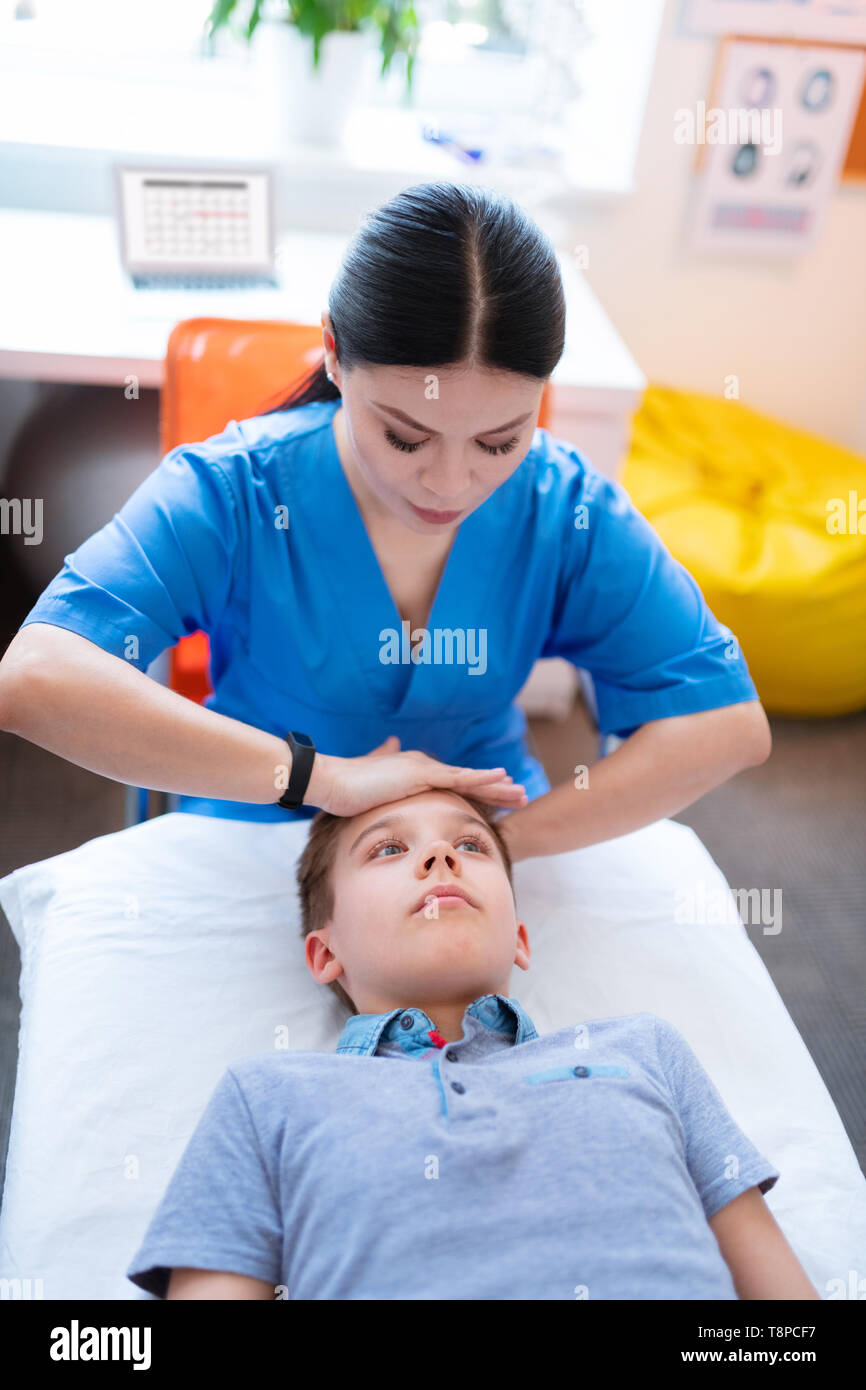 Erfahrene sanfte dunkelhaarige Frau in der blauen Uniform Drücken auf die Stirn Stockfoto
