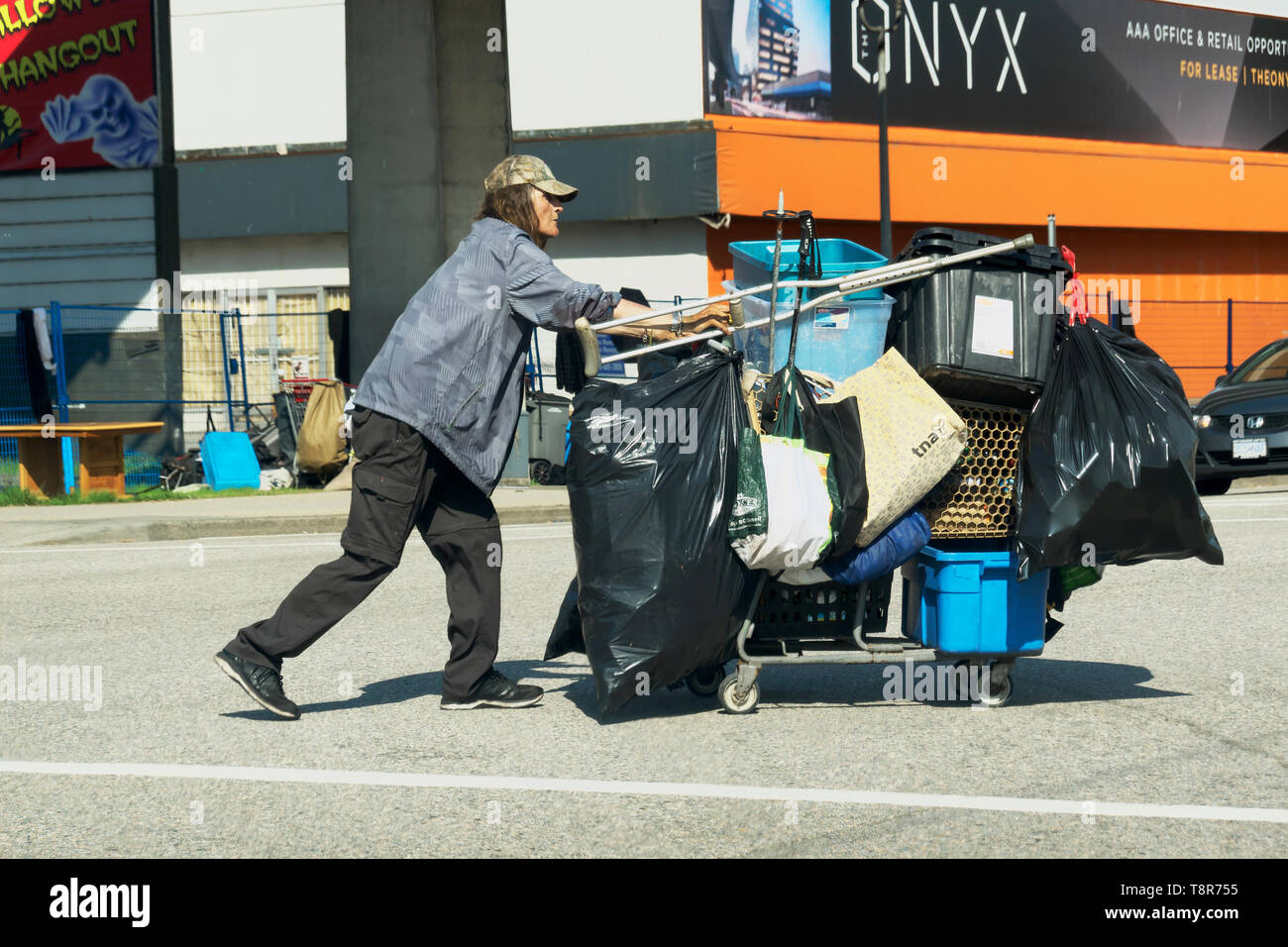 Ein obdachloser Mann gehen über eine Kreuzung treibt ein Warenkorb mit seinen weltlichen Besitz geladen. Stockfoto