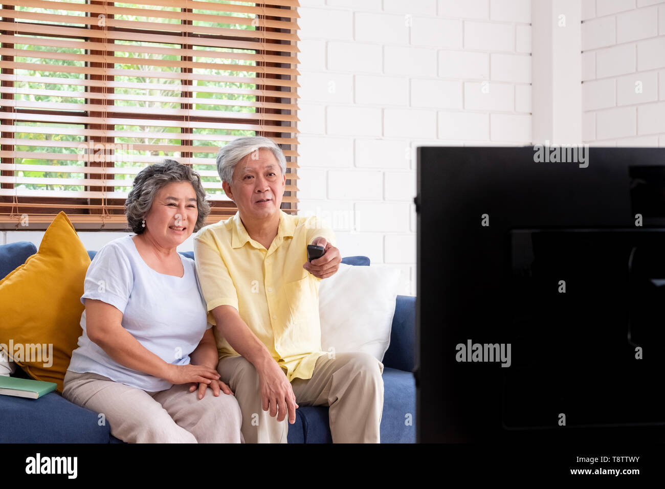 Asiatische paar Senior sitzen auf dem Sofa und die Fernbedienung verwenden, um den Kanal zu wechseln und Fernsehen im Wohnzimmer zu Hause. Panning von Blur Vordergrund telev Stockfoto