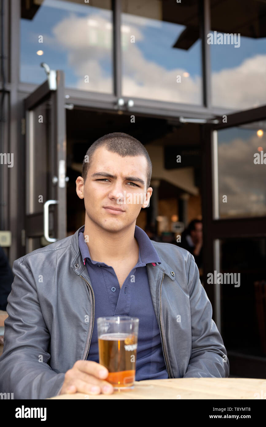 Porträt eines jungen Mannes mit Lederjacke, der in die Kamera schaut und ein Glas Bier auf der Außenterrasse hält. Junger Mann, der ein Glas Bier isst. Stockfoto