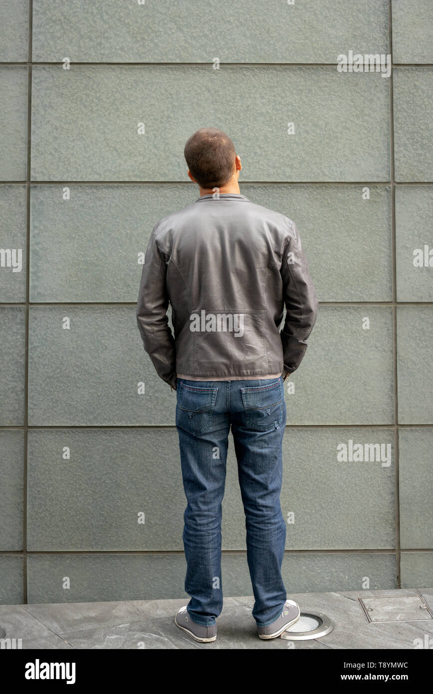 Junger 20-30-jähriger Mann in voller Größe mit Lederjacke und Jeans, der still steht und auf eine Wand blickt Stockfoto