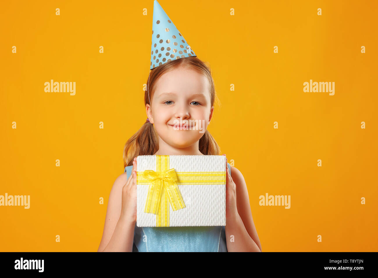 Fröhliches kleines Mädchen feiert Geburtstag. Das Kind hält einen Kasten mit einem Geschenk. Eine Party. Closeup Portrait auf gelben Hintergrund. Stockfoto