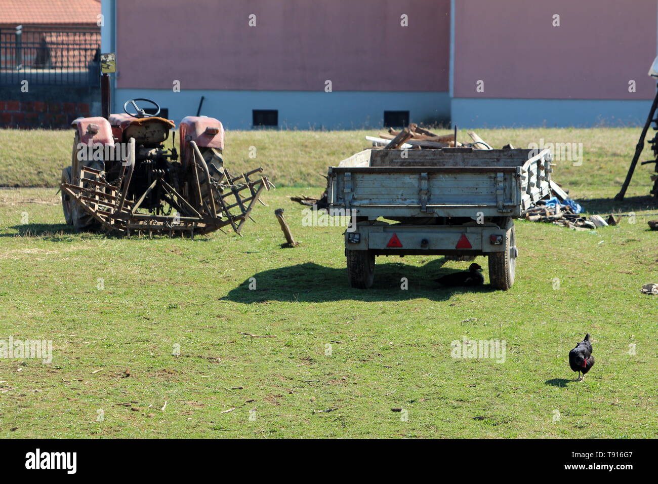 Retro Vintage alten Traktor mit angehängtem verrostete landwirtschaftliche Geräte neben Traktoren Anhänger links im Hinterhof mit ungeschnittenem Gras umgeben Stockfoto