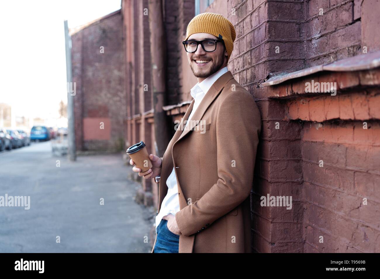 Hübscher junger Mann in Freizeitkleidung holding Einweg Becher und lächelnd während zu Fuß durch die Stadt. Stockfoto