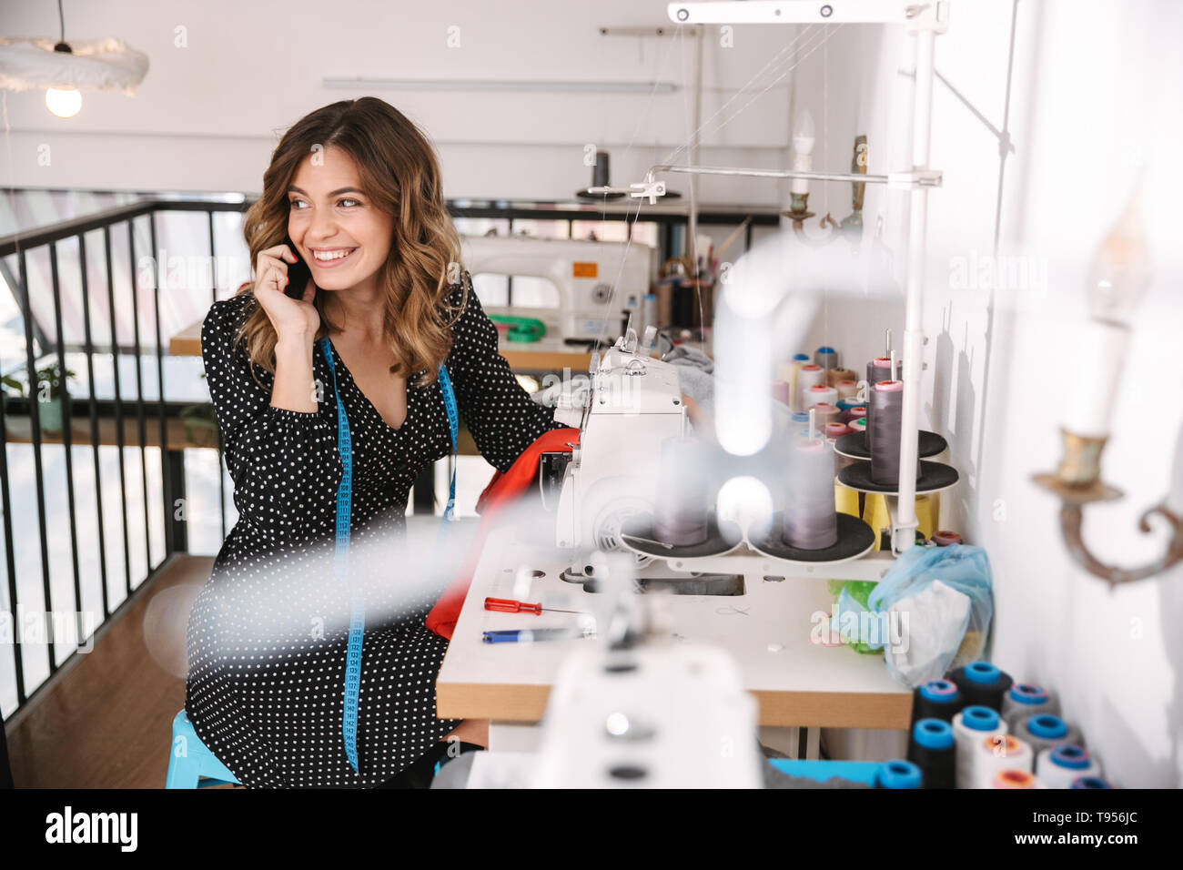 Lächelnden jungen Frau Näherin in der Werkstatt arbeiten, mit Nähmaschine, Gespräch am Handy Stockfoto