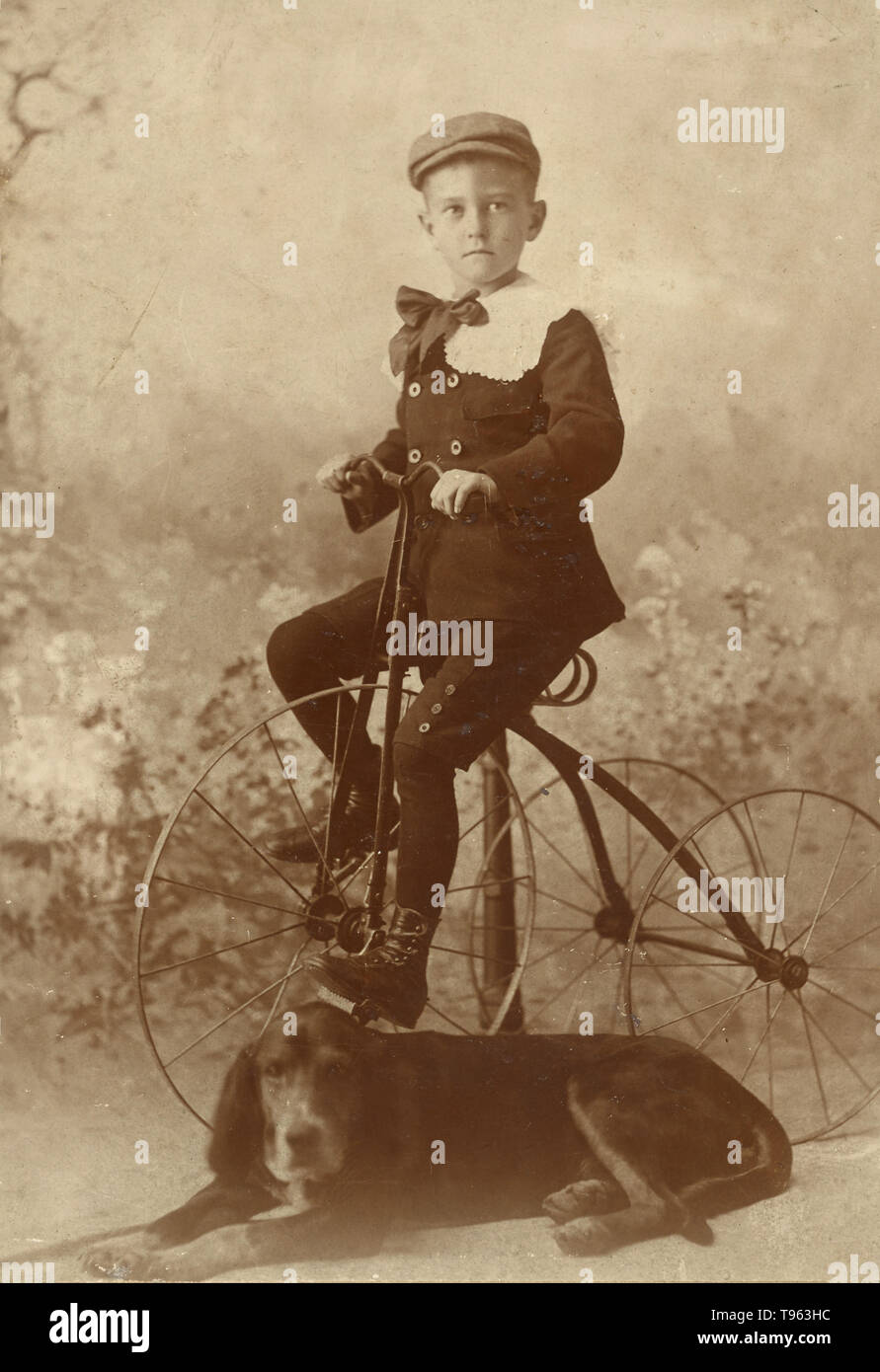 Junge trägt eine Kappe, montiert auf einem alten Stil dreirädrigen Fahrrad, mit einem Hund im Vordergrund, 1890. Von Henry Davis, Amerikanische genommen. Stockfoto