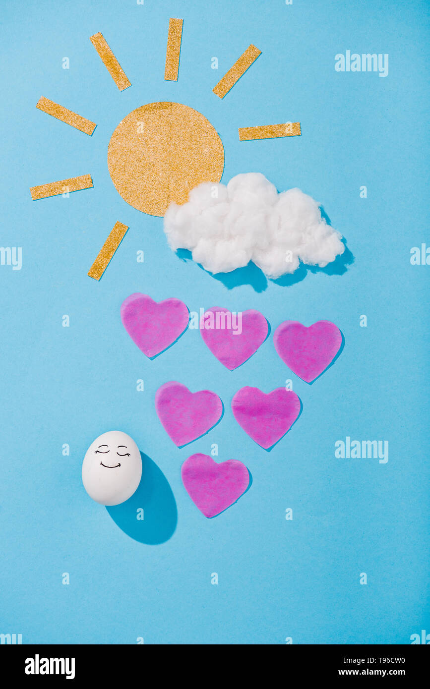 Blick von oben auf die Papier sun, Zuckerwatte Cloud, Ei mit glücklichen Gesichtsausdruck und Herzförmigen Regentropfen auf Blau Stockfoto