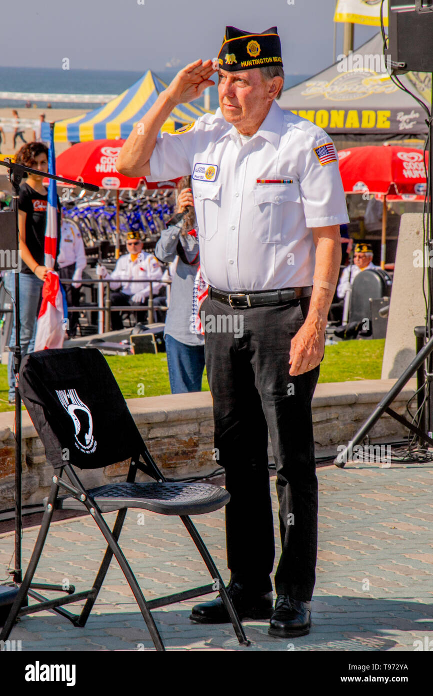 Eine militärische Veteran Mitglied der amerikanischen Legion begrüßt nach dem POW/MIA-Flag auf einen symbolischen leeren Stuhl mit einem Sinnbildlichen T Shirt zu einem Veterans Day Einhaltung in Huntington Beach, CA. Stockfoto