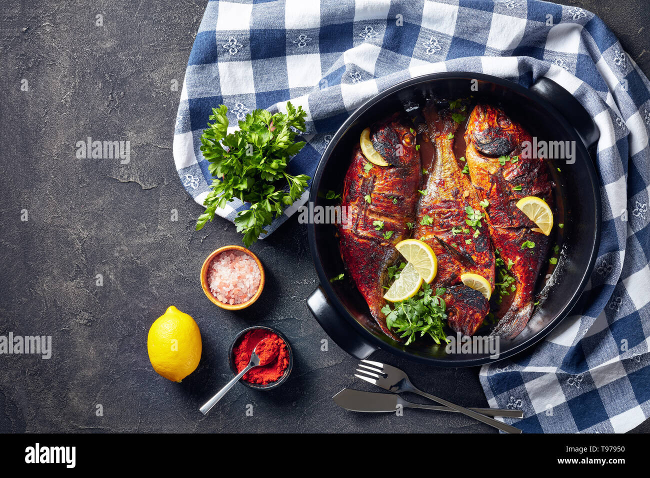 Köstliche, frisch gegrillte dorado Fisch in einem schwarzen Keramik Auflaufform auf einer konkreten Tabelle mit einem Küchentuch und Petersilie, Ansicht von oben, flatlay Stockfoto