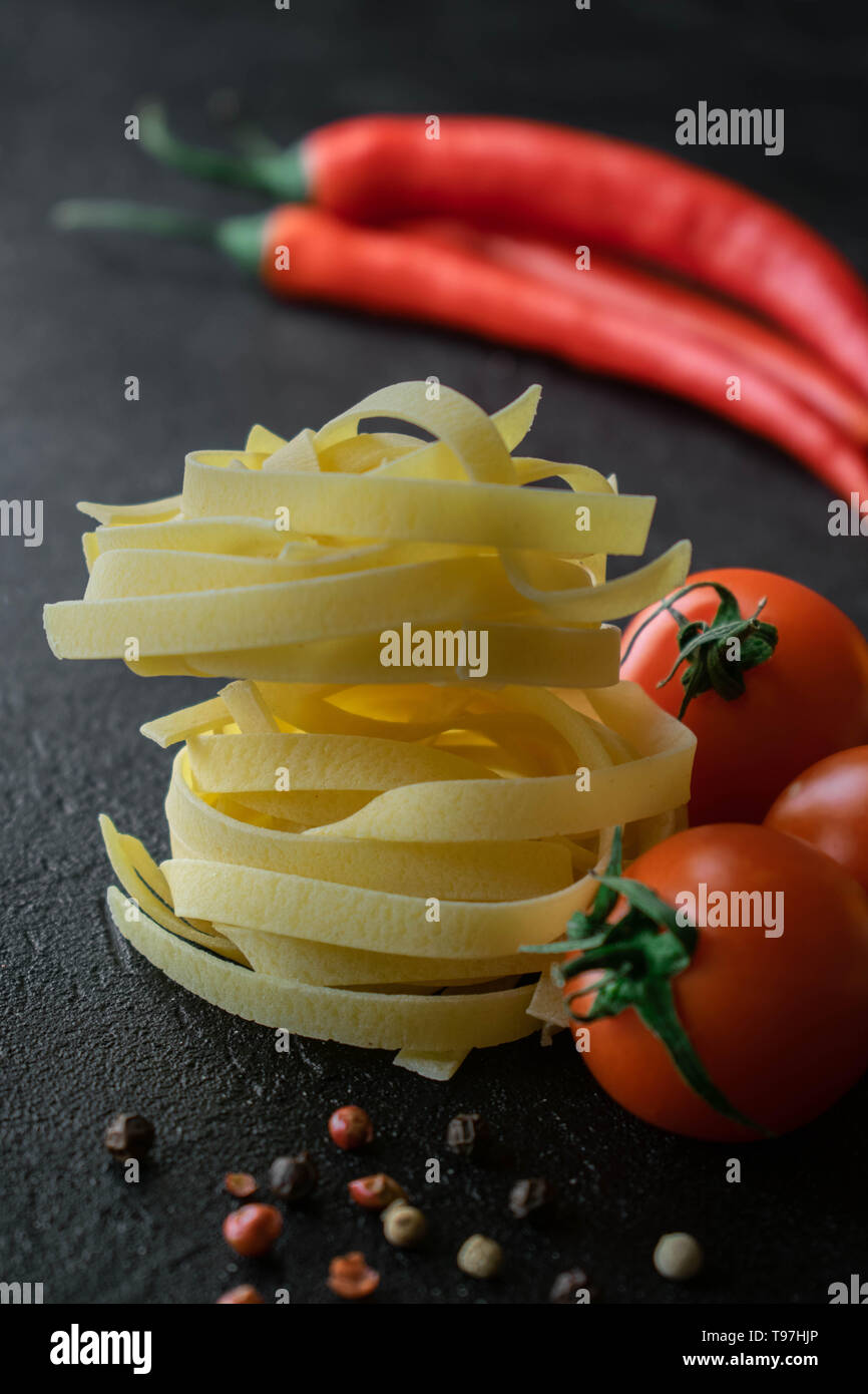 Auf schwarzem Hintergrund Kugeln von Roh Fettuccine Pasta mit frischen Tomaten mit Wurzeln, schwarze und rote Pfefferkörner und zwei Kaffeepads von Chili Stockfoto