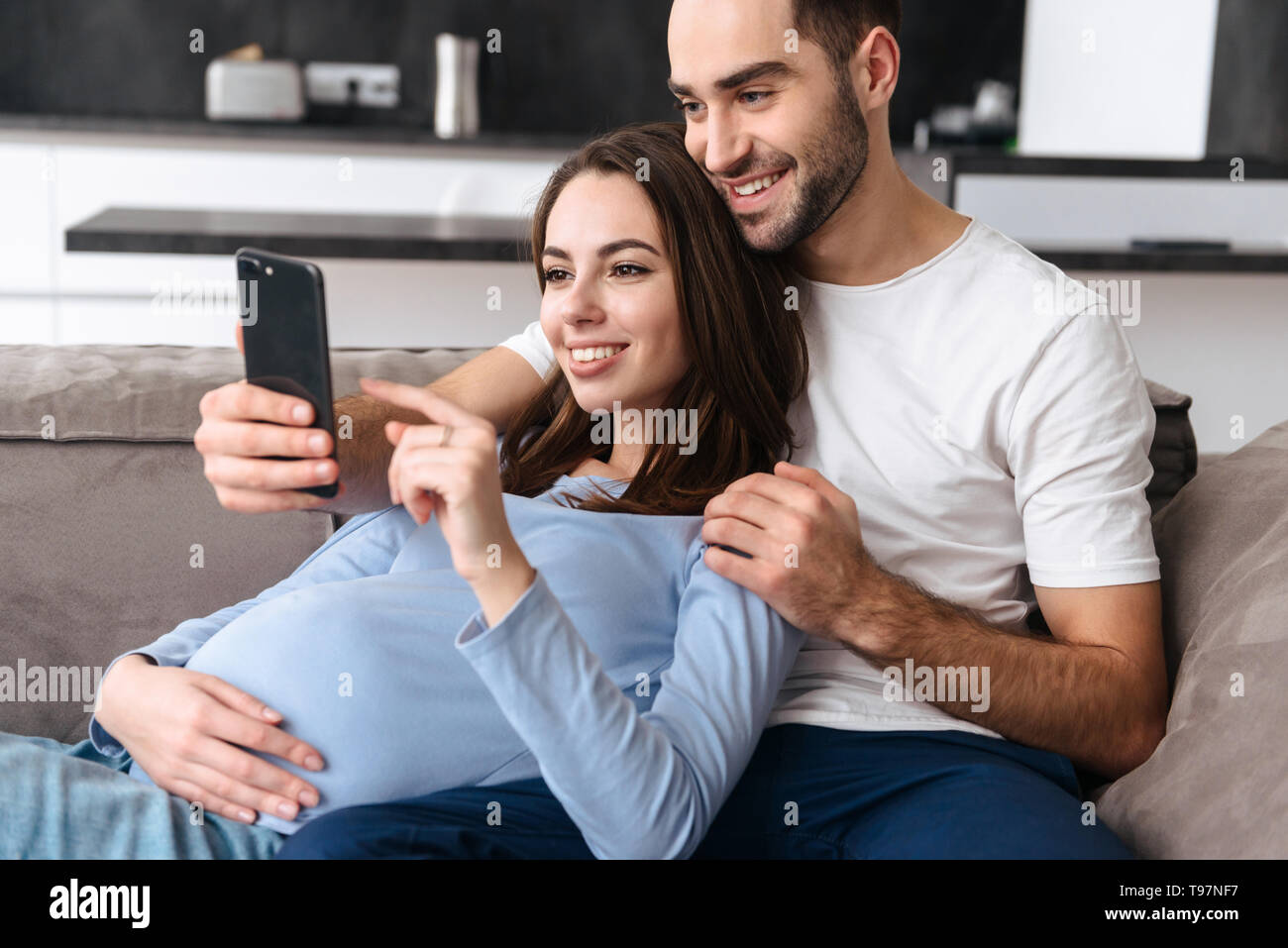 Bild der Europäischen schwangere Frau und gutaussehenden Mann in Freizeitkleidung liegend auf dem Sofa im Wohnzimmer zu Hause und Sie ihr Smartphone Stockfoto
