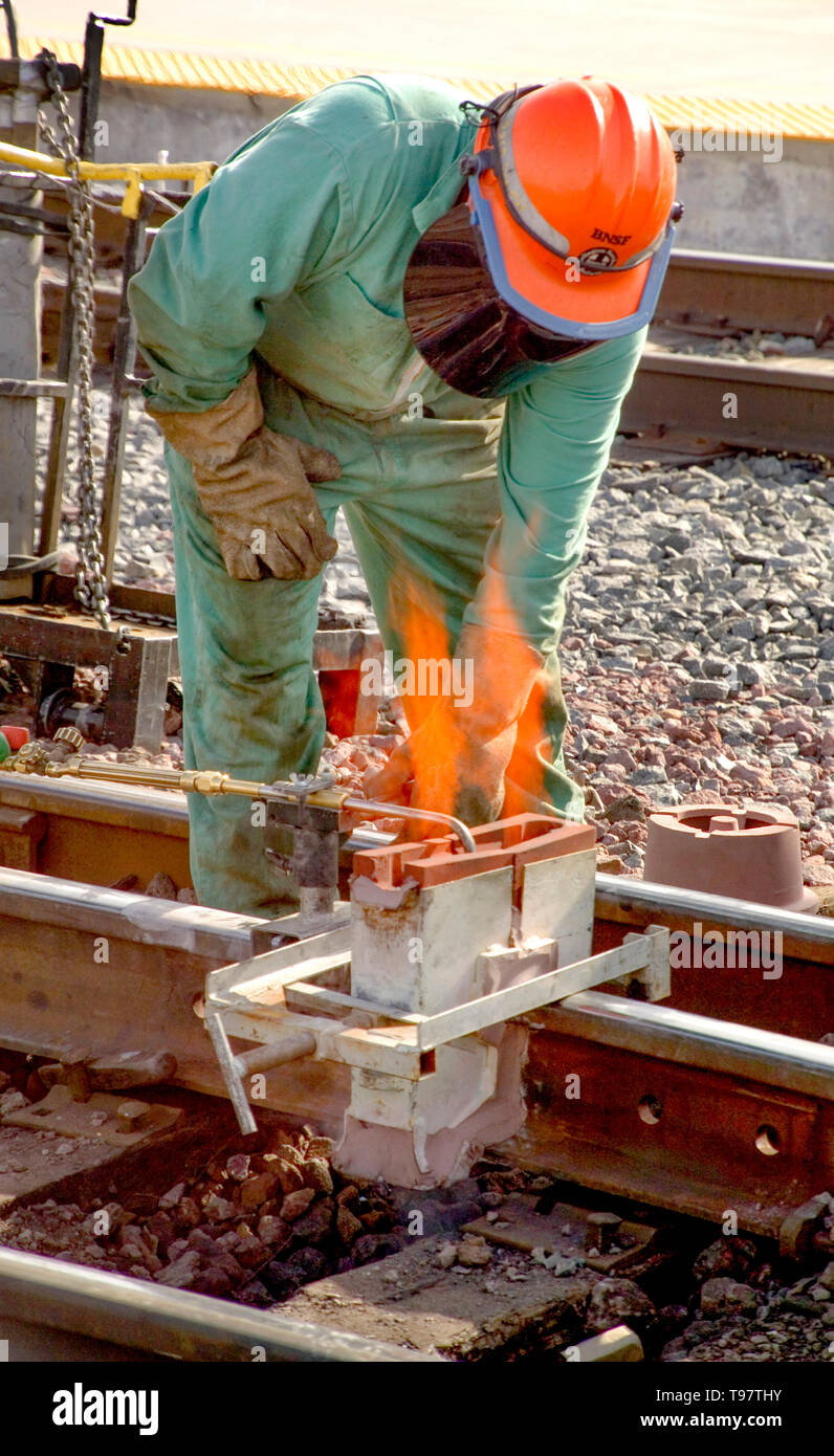 Trägt einen Schutzhelm und Gesichts Schutzmaske, ein Zug Wartung Arbeiter beaufsichtigt eine Eisenbahnschiene Vorgehensweise zur Verklebung mit Metallschmelze in der San Bernardino, CA, Zug Yards. Stockfoto