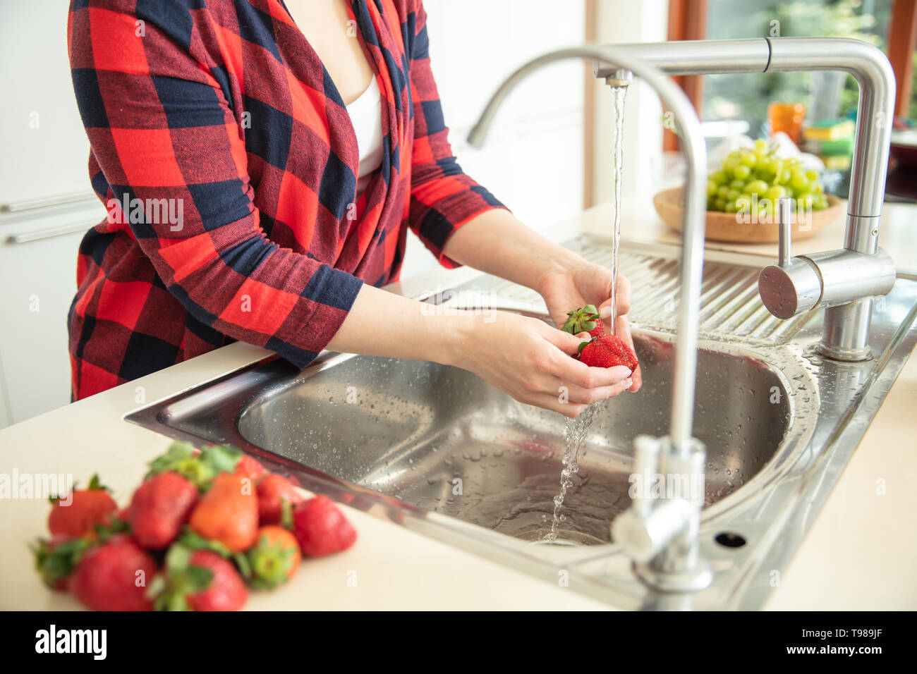 Frau spült die Erdbeeren in der Küche mit grünen Trauben im Hintergrund und roter Erdbeeren auf den ersten Plan. Stockfoto