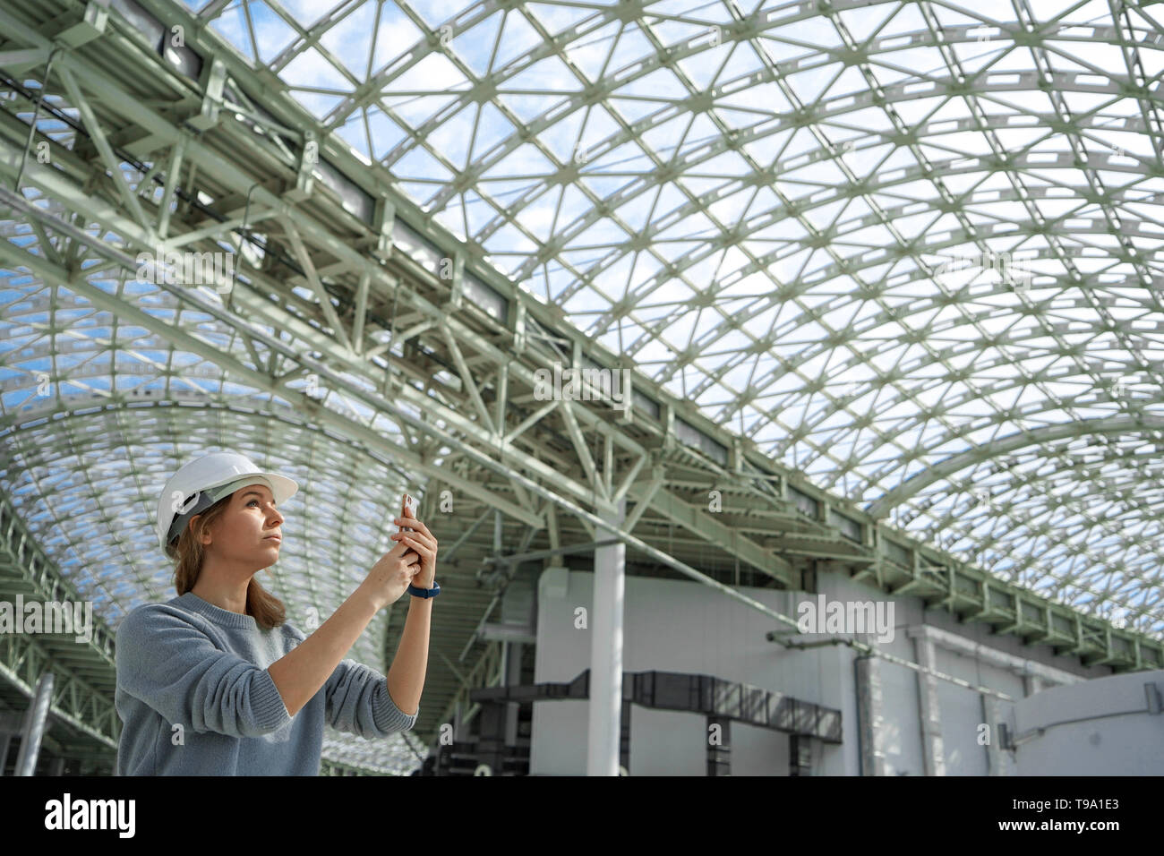 Ein junges Mädchen in einen Helm in einem großen Zimmer mit einem Glasdach nimmt Bilder auf dem Handy. Eine ernsthafte Experte, eine Frau das Objekt kontrolliert Stockfoto