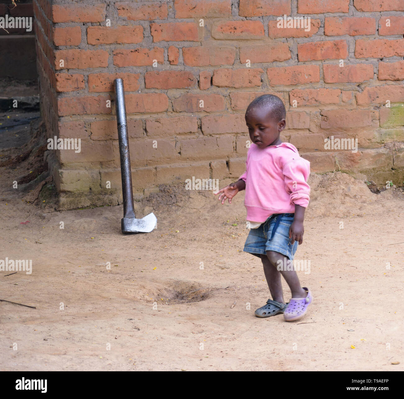 Eine malawische Kind geht vor einem großen Metall ax Linke lehnte sich gegen eine Wand nach dem Zerkleinern von Brennholz Stockfoto