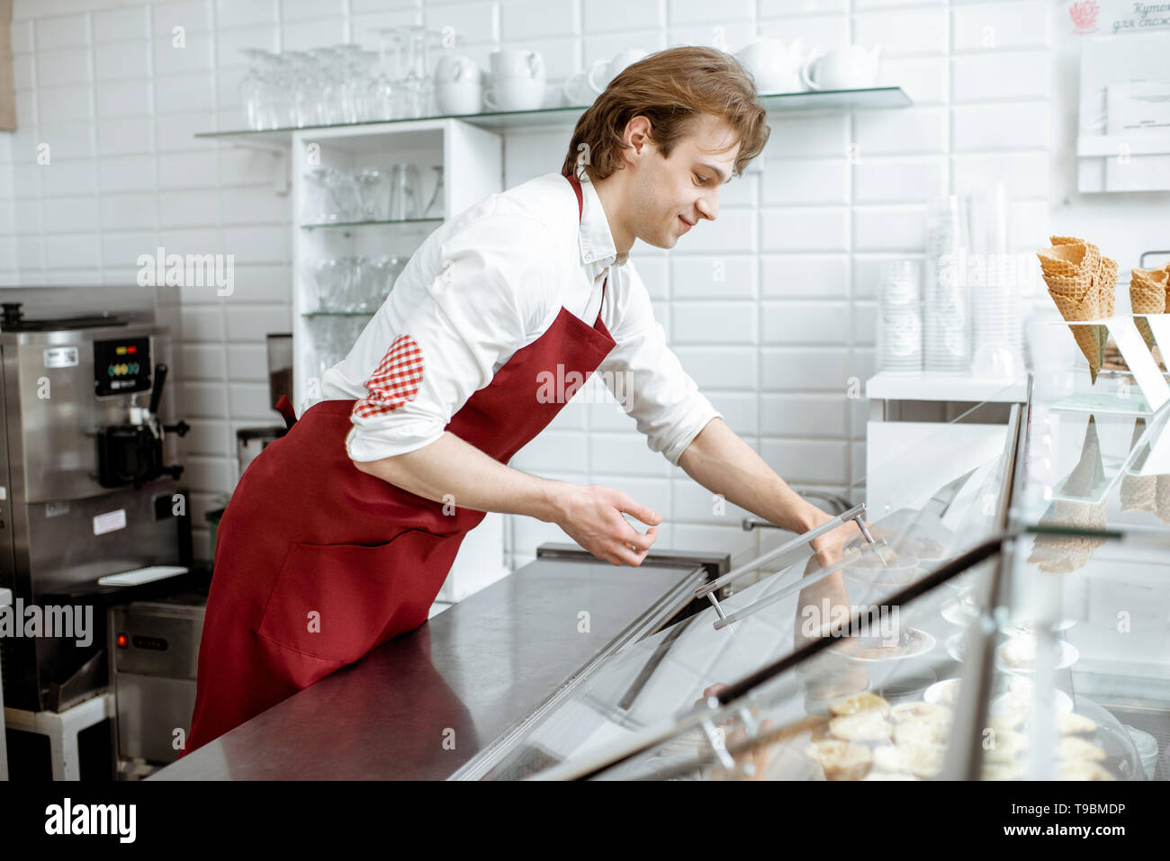 Junge Verkäufer in rot Schürze, süßes Gebäck im Kühlschrank des Showcase an der modernen Konditorei und Café Stockfoto