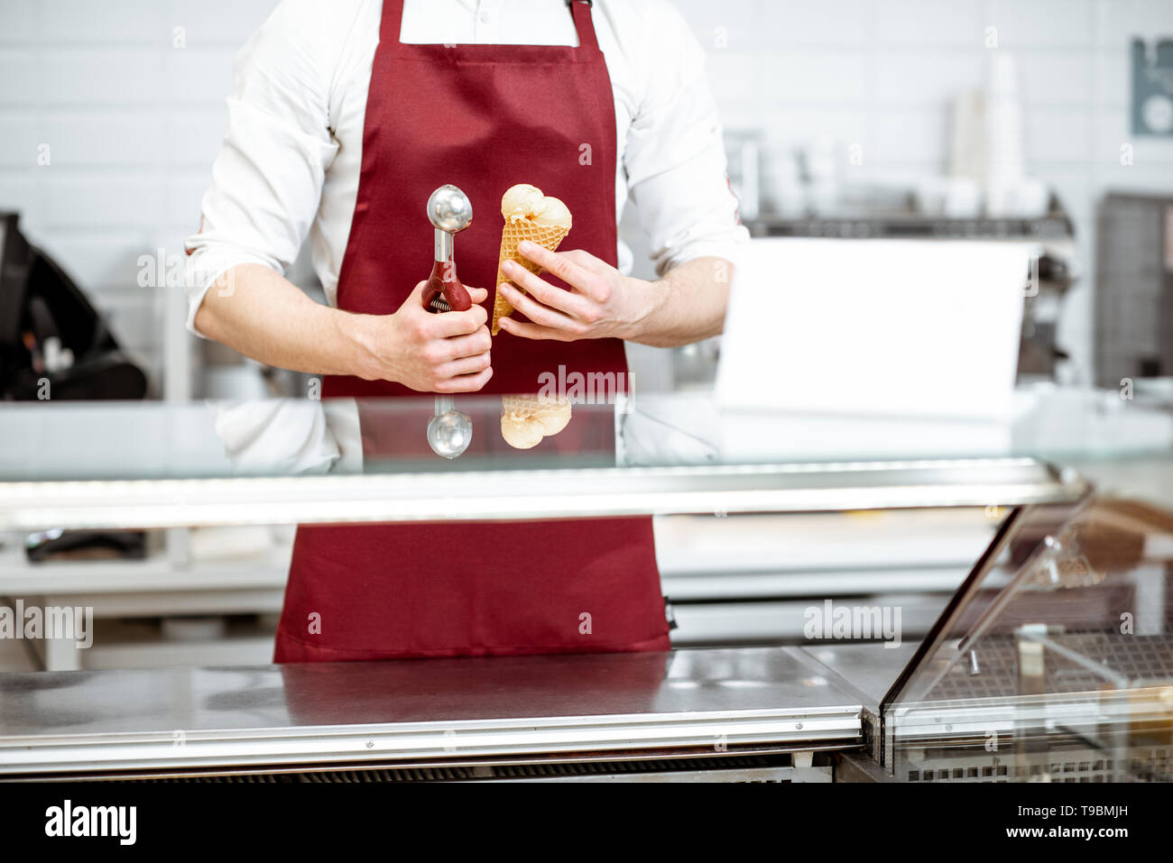 Verkäufer halten Eis in der Waffel Kegel und professionelle Schaufel auf die rote Schürze, Hintergrund, Nahaufnahme Stockfoto